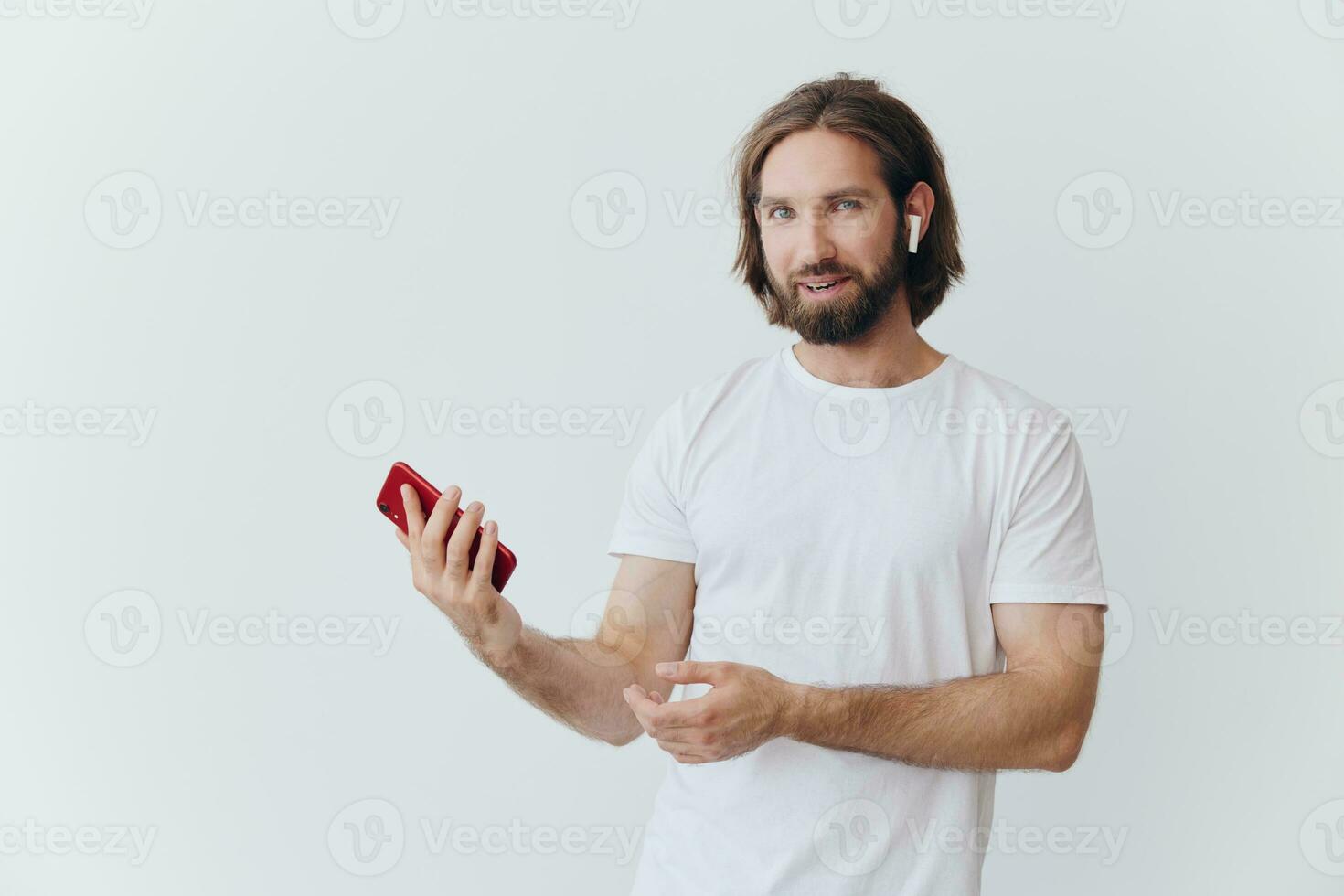 A man with a hipster beard in a white T-shirt with a phone and wireless headphones smiling listening to music and an audiobook online against a white wall photo