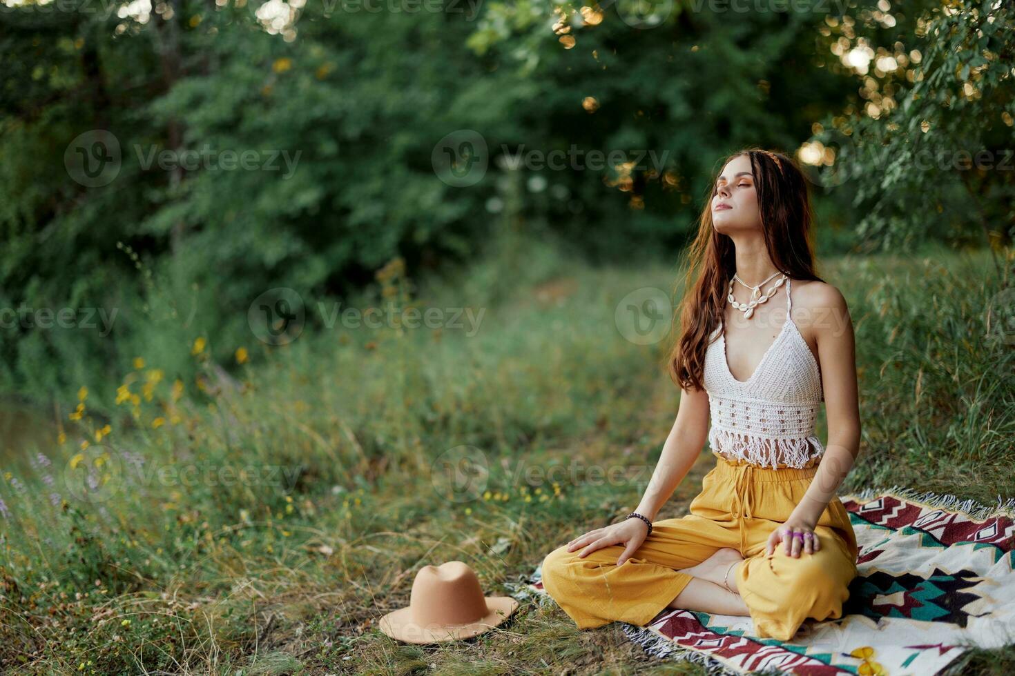 A young hippie woman meditates in nature in the park, sitting in a lotus position on her colorful plaid and enjoying harmony with the world in eco-clothing photo