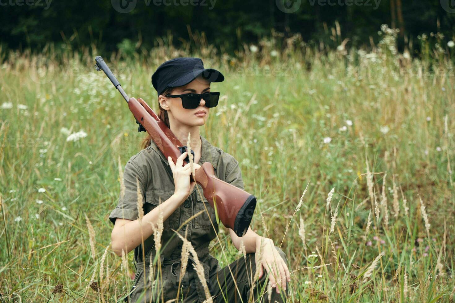 mujer en naturaleza se sienta en un abrigo con un arma oscuro lentes negro gorra verde foto