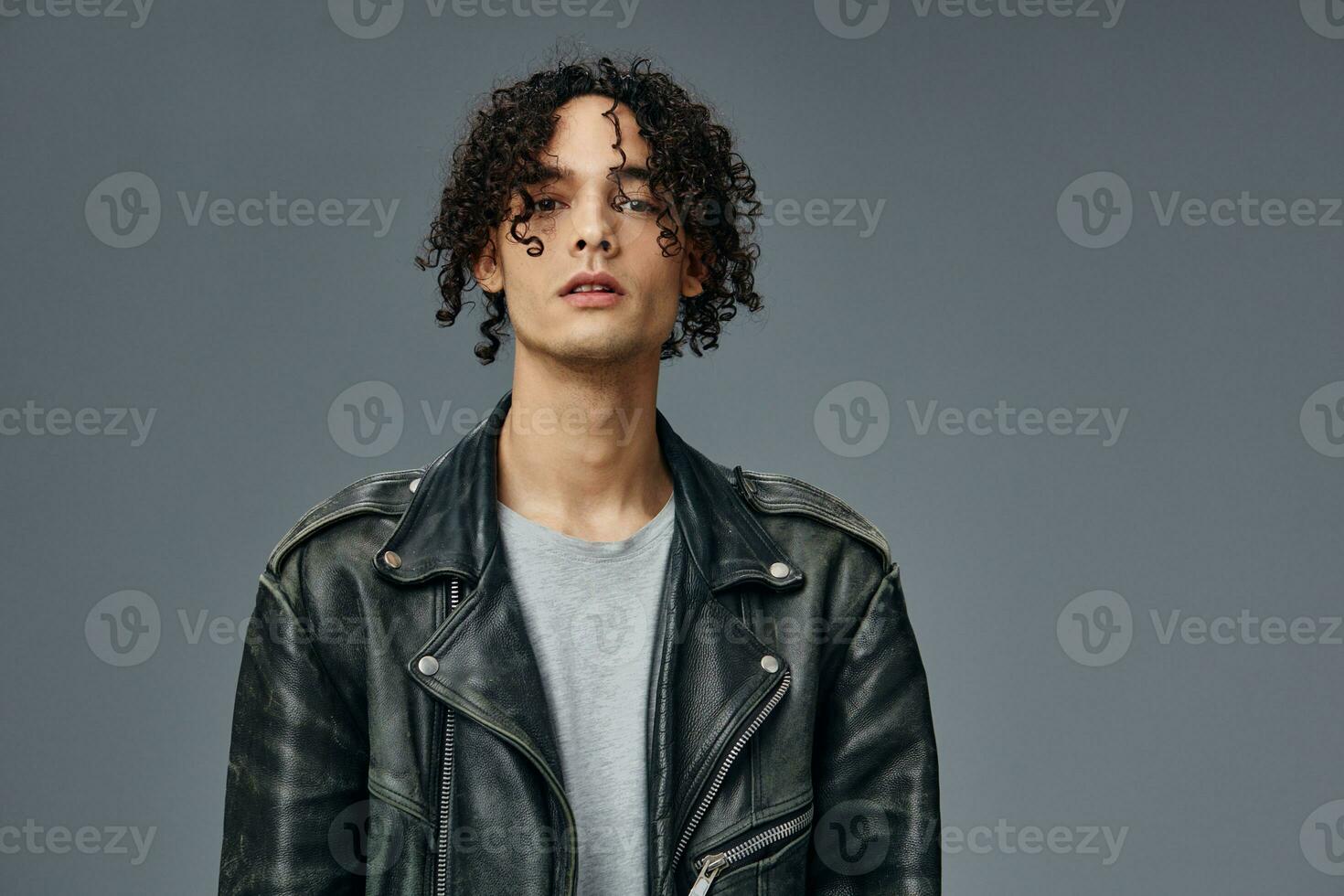 Portrait of serious stylish tanned curly man leather jacket looks at camera posing isolated on gray studio background. Cool fashion offer. Huge Seasonal Sale New Collection concept. Copy space for ad photo