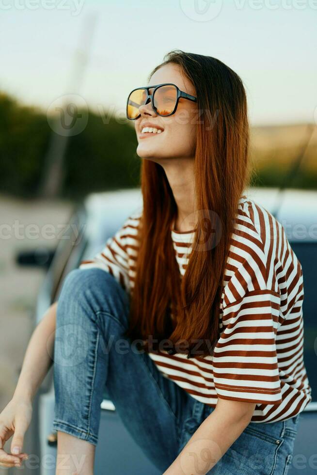 A fashionable woman smiles sweetly in stylish sunglasses, a striped t-shirt and jeans, sits on the trunk of a car and looks at the beautiful nature of autumn. Travel lifestyle photo