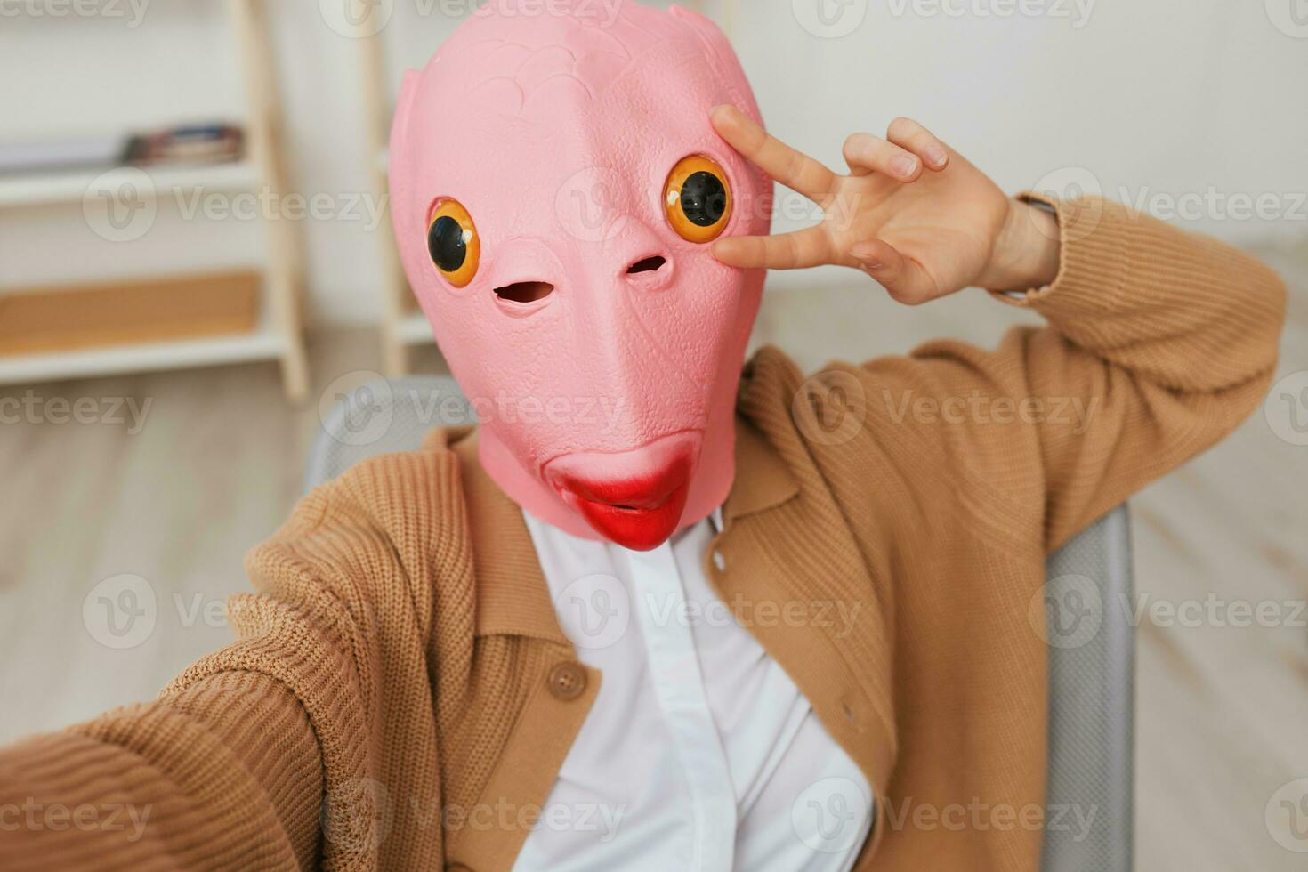Funny blonde lady in pink fish mask warm sweater doing selfie use show v sign gesture phone in armchair at modern home. Pause from work, take a break, social media in free time concept. Wide angle photo