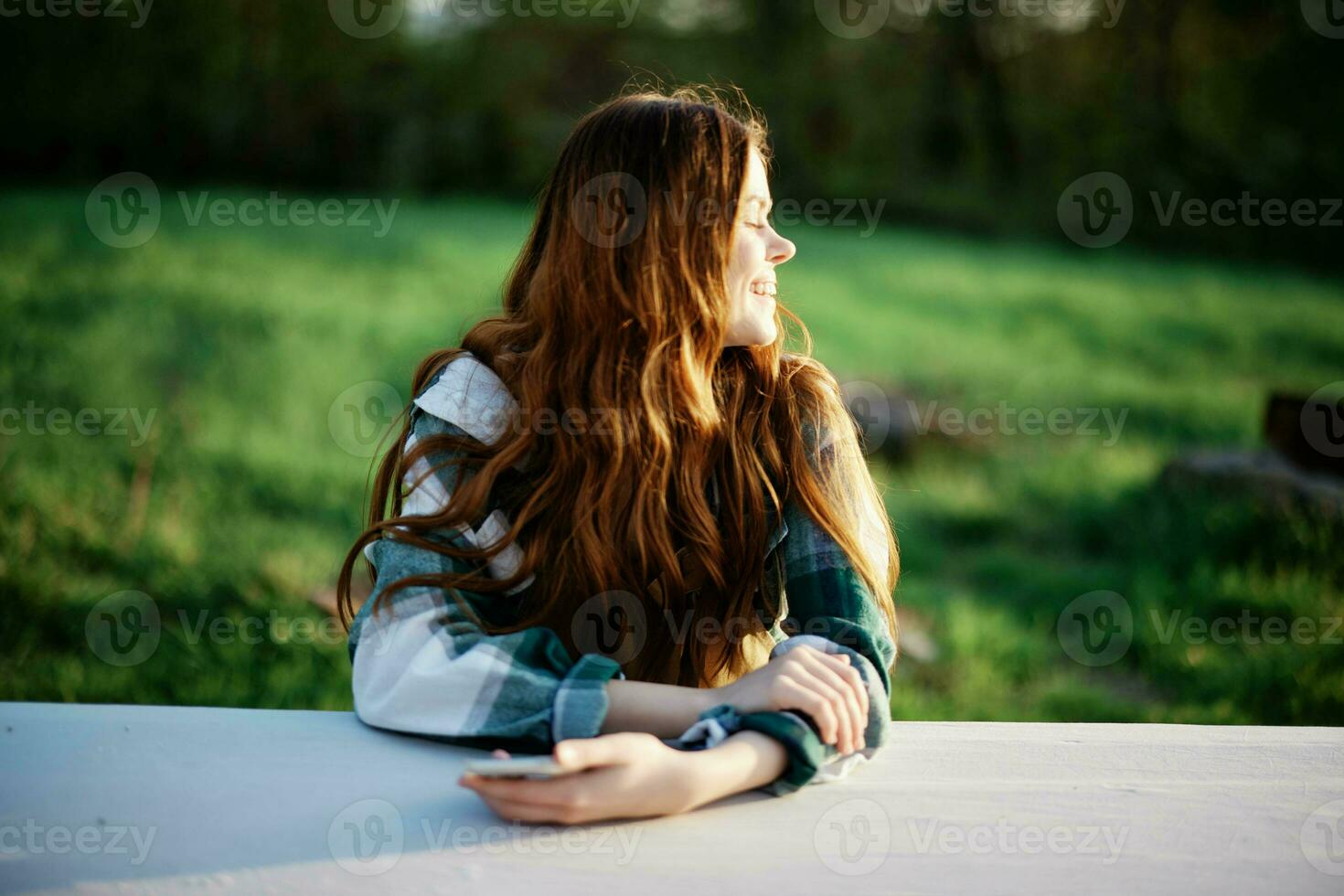 Girl with phone in hand sitting in nature in the park at the table smiling beautifully and looking at the camera with her red hair lit by the sunset sunlight of summer photo