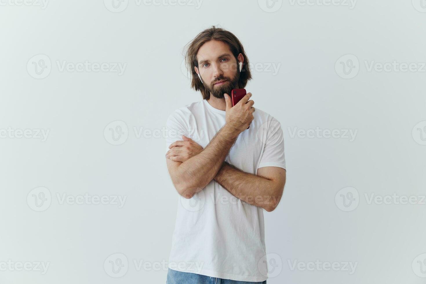 A man with a beard and long hair in a white T-shirt and blue jeans stands against a white wall, leaning against it and listening to music with wireless white headphones, staring thoughtfully photo