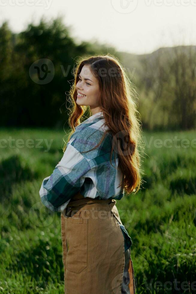 A woman gardener in an apron stands in a field of green grass outdoors, smiling on a summer day into a sunset photo