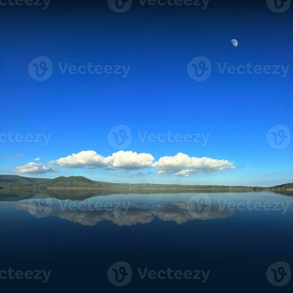 Reflection of clouds on the lake with half moon in the blu sky photo