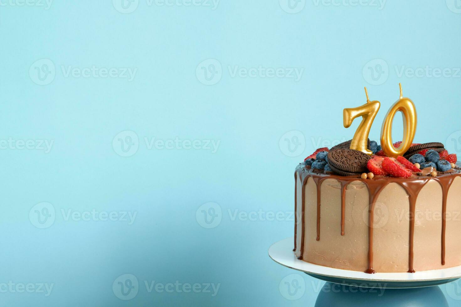 Chocolate birthday cake with berries, cookies and number seventy golden candles on blue wall background, copy space photo