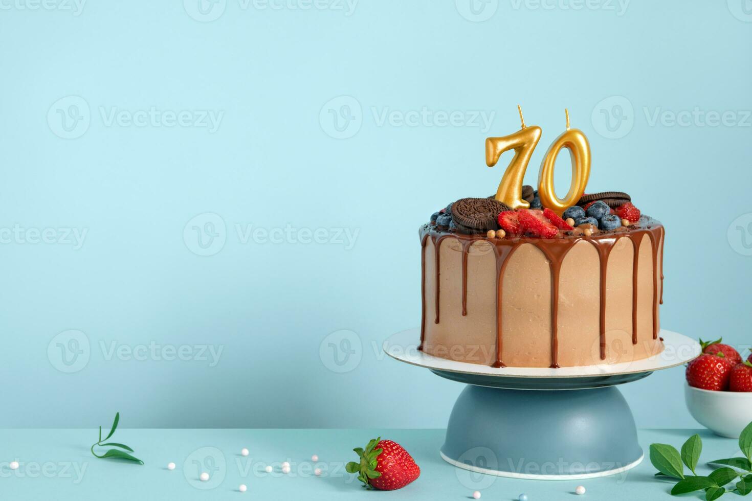 Chocolate birthday cake with berries, cookies and number seventy golden candles on blue wall background, copy space photo