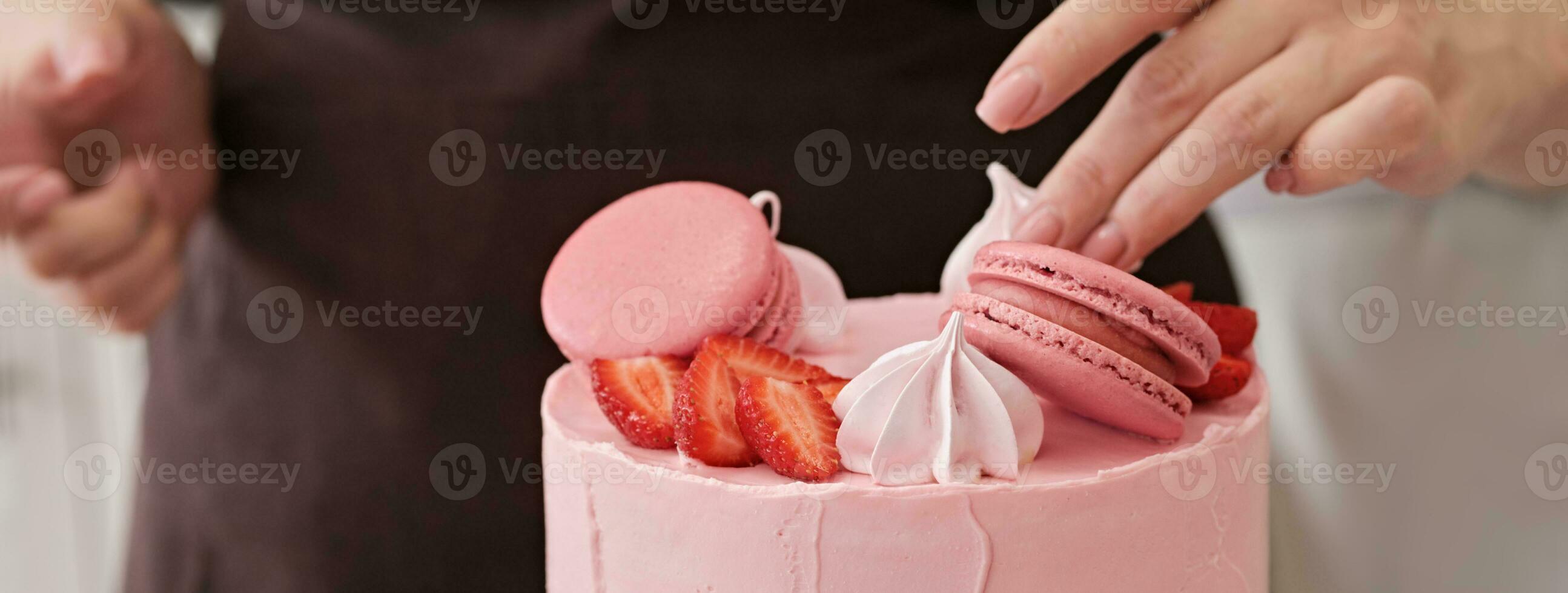 Woman pastry chef decorates pink cake with macaroons and berries, close-up. Cake making process, Selective focus. Banner format photo