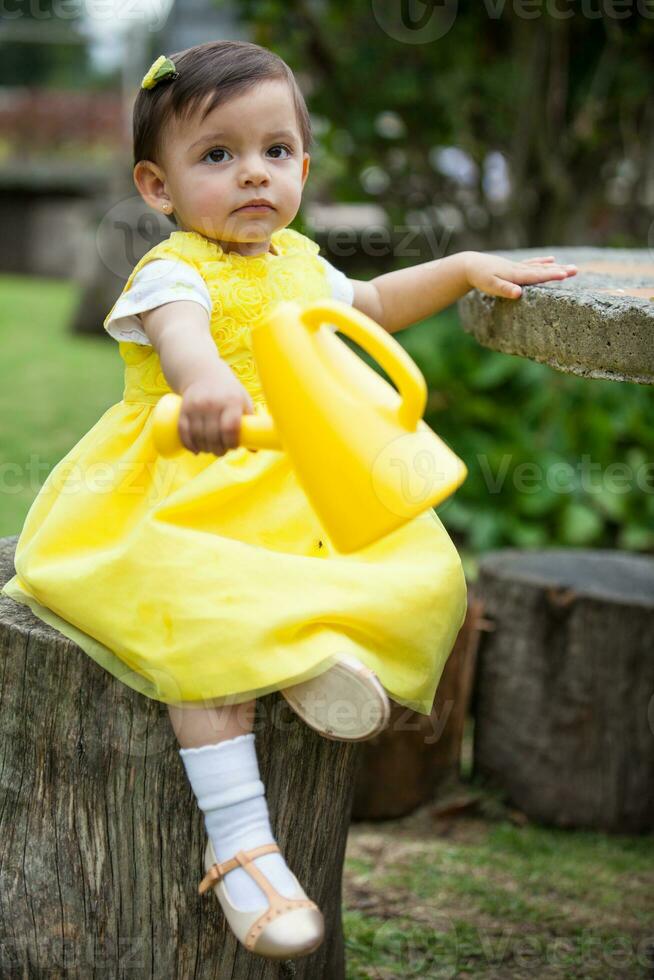 dulce uno año antiguo bebé niña vestido en amarillo riego el plantas a el jardín foto