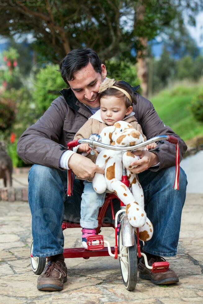 joven padre jugando con su uno año antiguo bebé al aire libre. con papá montando mi triciclo foto