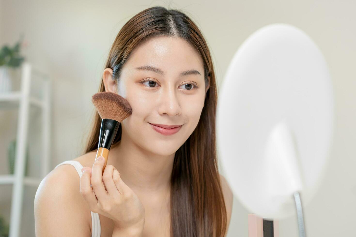 Happy beauty blogger concept, cute asian young woman, girl smile, make up face by applying brush blush powder on her cheek, looking at the mirror. People look with natural fashion style. photo