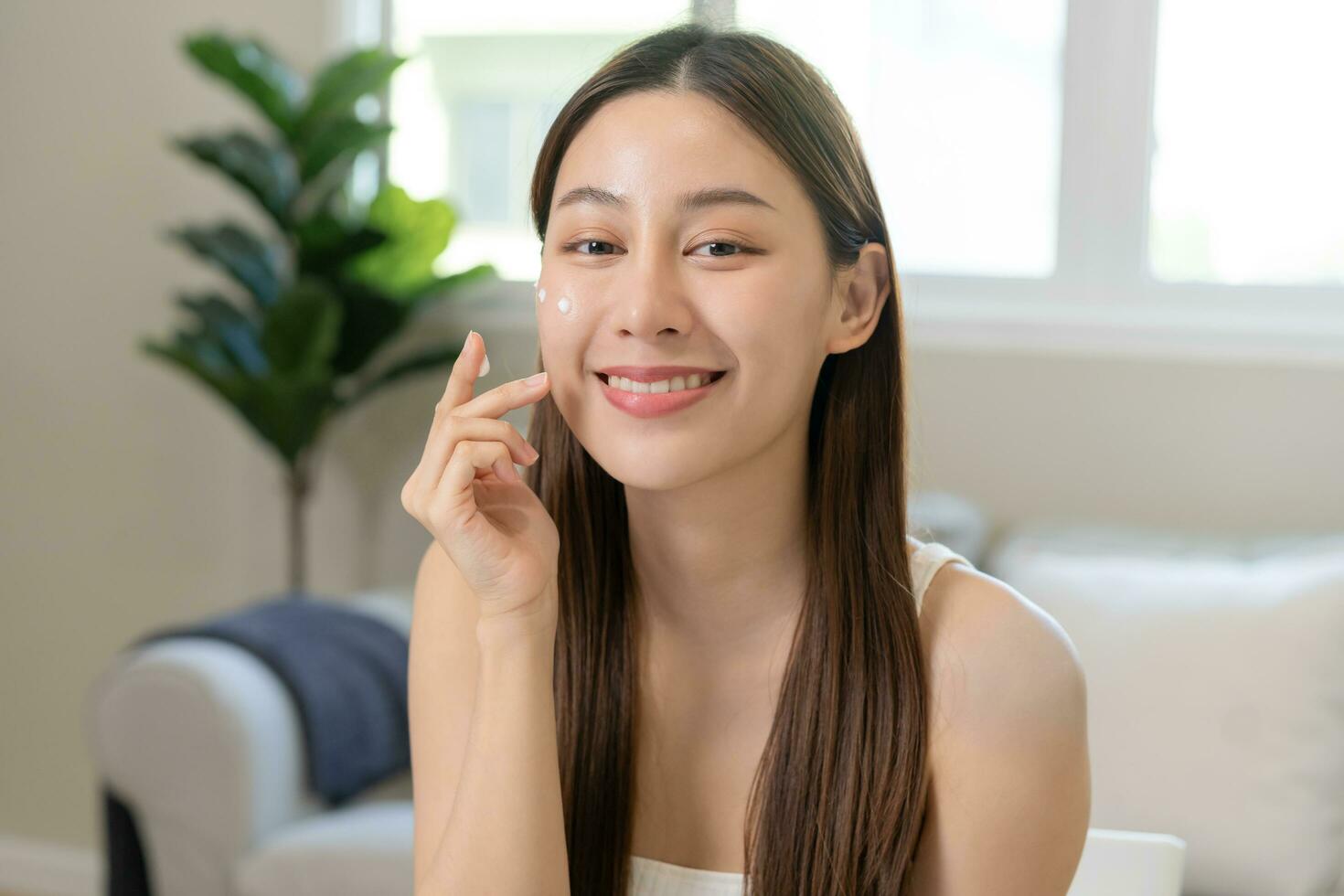 Fresh healthy skin, beautiful smile of asian young woman, girl looking at mirror, applying moisturizer on her face, putting cream treatment before makeup cosmetic routine at home. Facial Beauty. photo