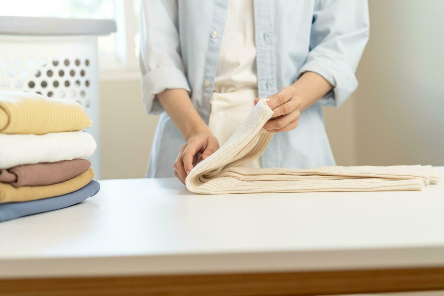 ama de casa, asiático joven mujer mano en muchos plegable recién camisas, suéteres o vestir en escritorio, mesa después Lavado limpiar ropa y el secado, haciendo casa trabajando en habitación a hogar. lavandería y mucama. foto