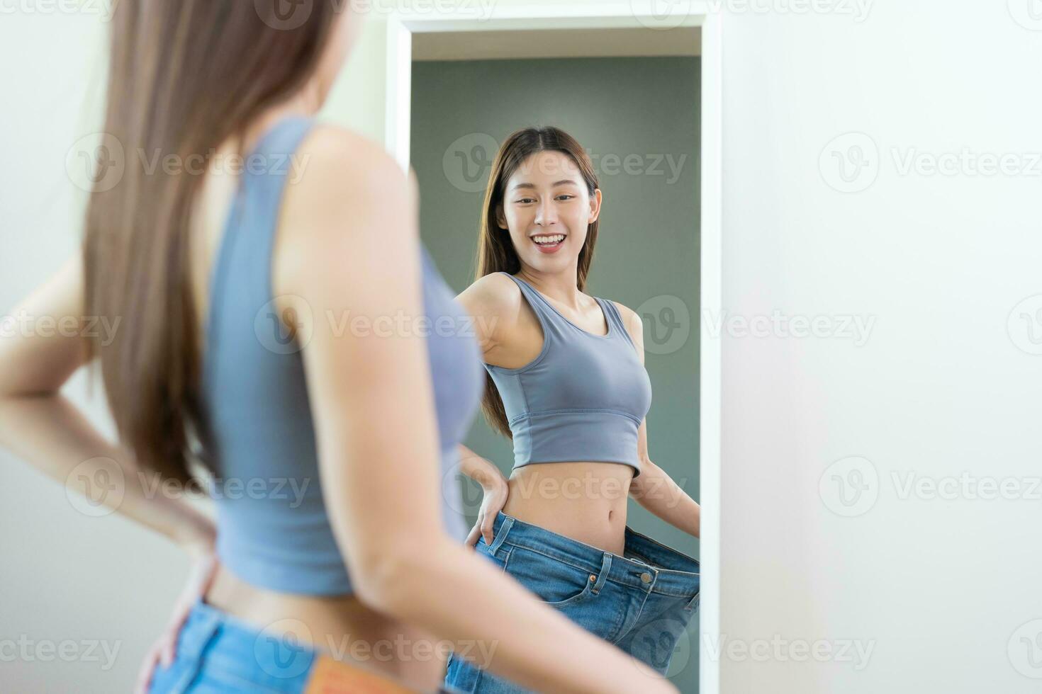 Shape slender, thin waist, attractive slim asian young woman, beautiful girl hand show shape her weight loss, wearing in big, large or over size jeans and looking into mirror. People body fit healthy. photo