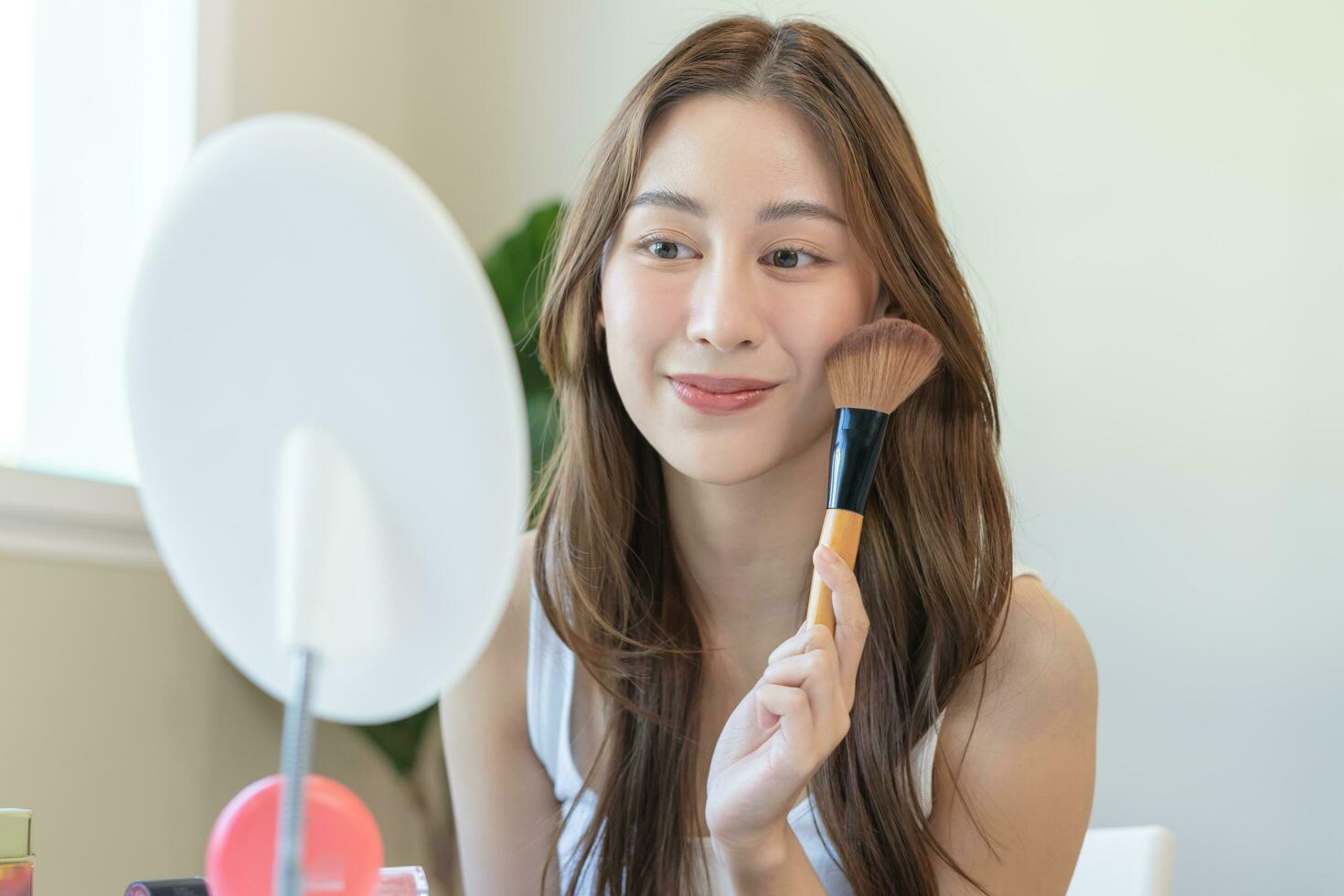 Happy routine beauty concept, pretty asian young woman, girl make up face by applying powder foundation by brush around face, looking at the mirror at home. Female look with natural fashion style. photo