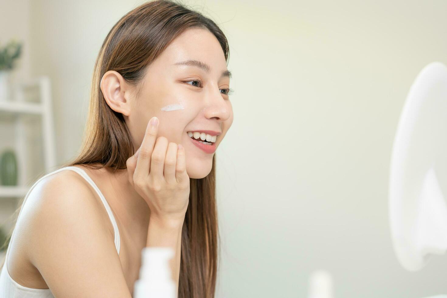 Fresh healthy skin, beautiful smile of asian young woman, girl looking at mirror, applying moisturizer on her face, putting cream treatment before makeup cosmetic routine at home. Facial Beauty. photo