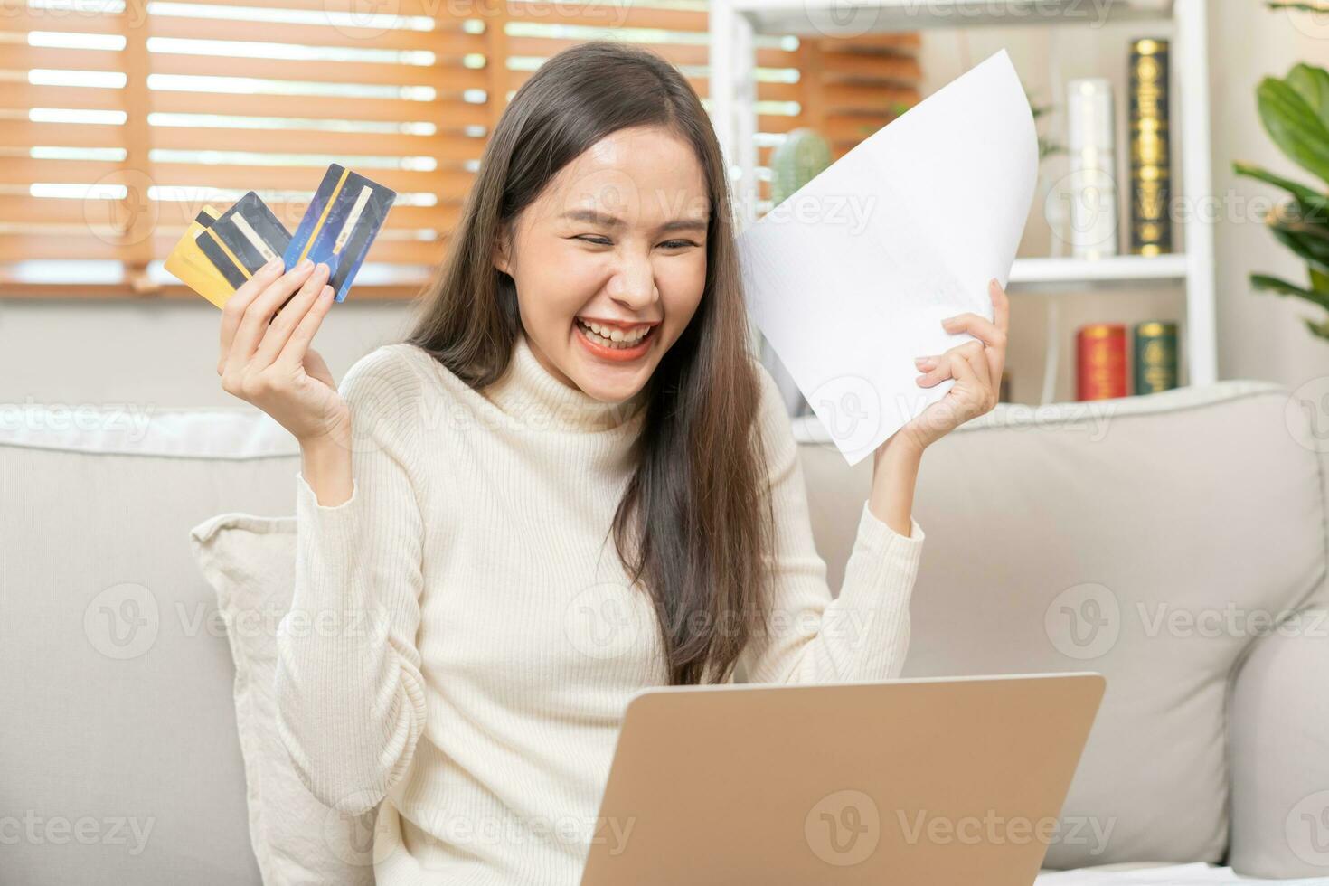 Happy excited, asian young woman, girl holding credit card and paperwork getting, received job promotion, approve tax refund cash back, looking good news at laptop computer, sitting on sofa at home. photo