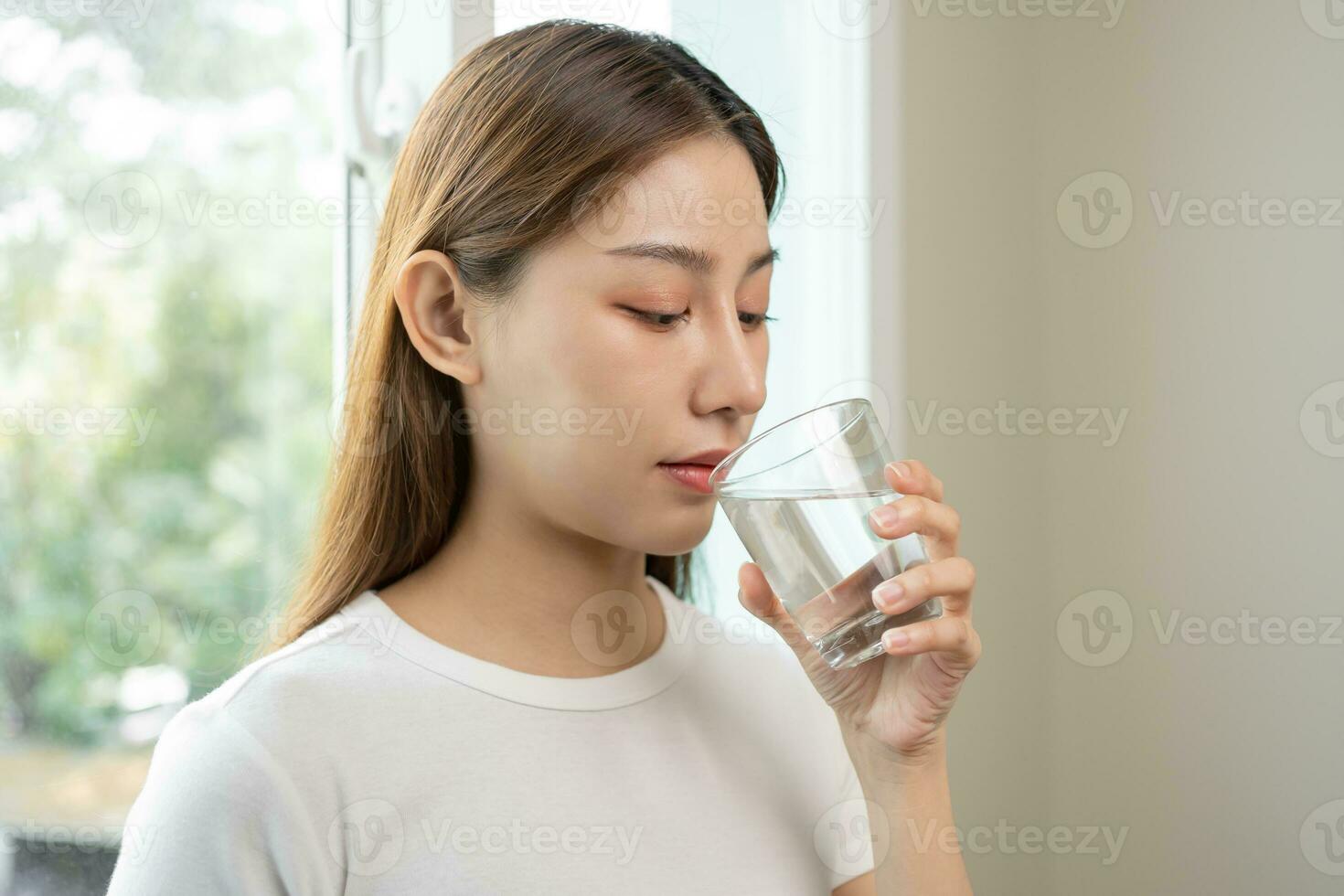 Happy beautiful, asian young woman, girl drinking, sip fresh glass of water for hydration of body, holding transparent glass in her hand, thirsty at home. Health care, healthy lifestyle concept. photo