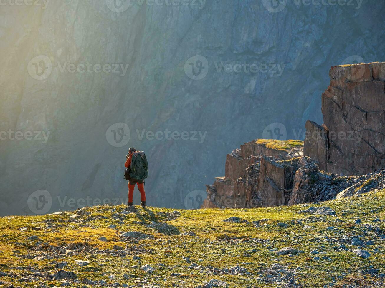 Photographer with a large backpack takes pictures of a beautiful mountain landscape on the edge of a cliff. photo