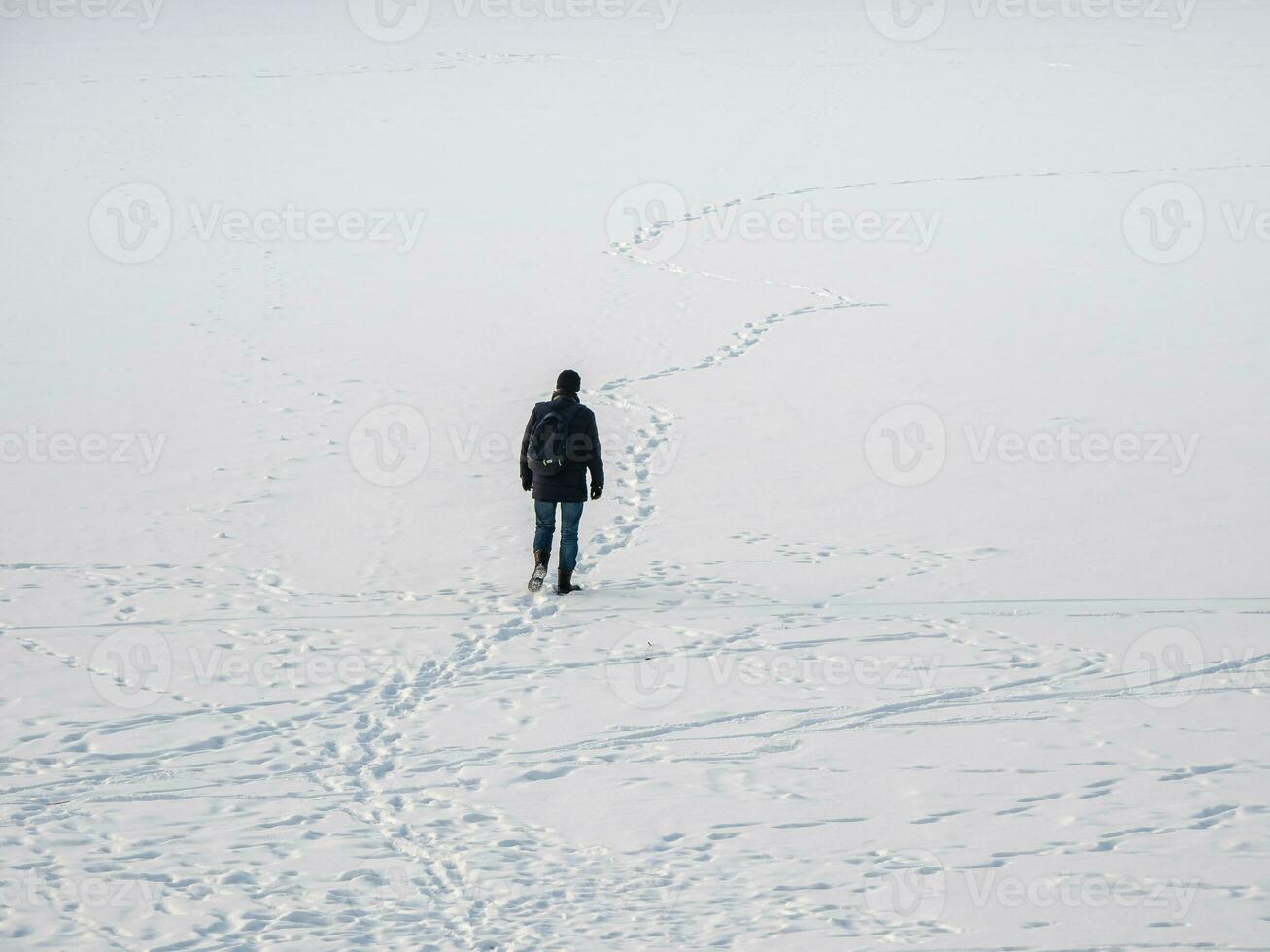 A man in a black jacket with a backpack walking on snow, footprints in snow, behind photo