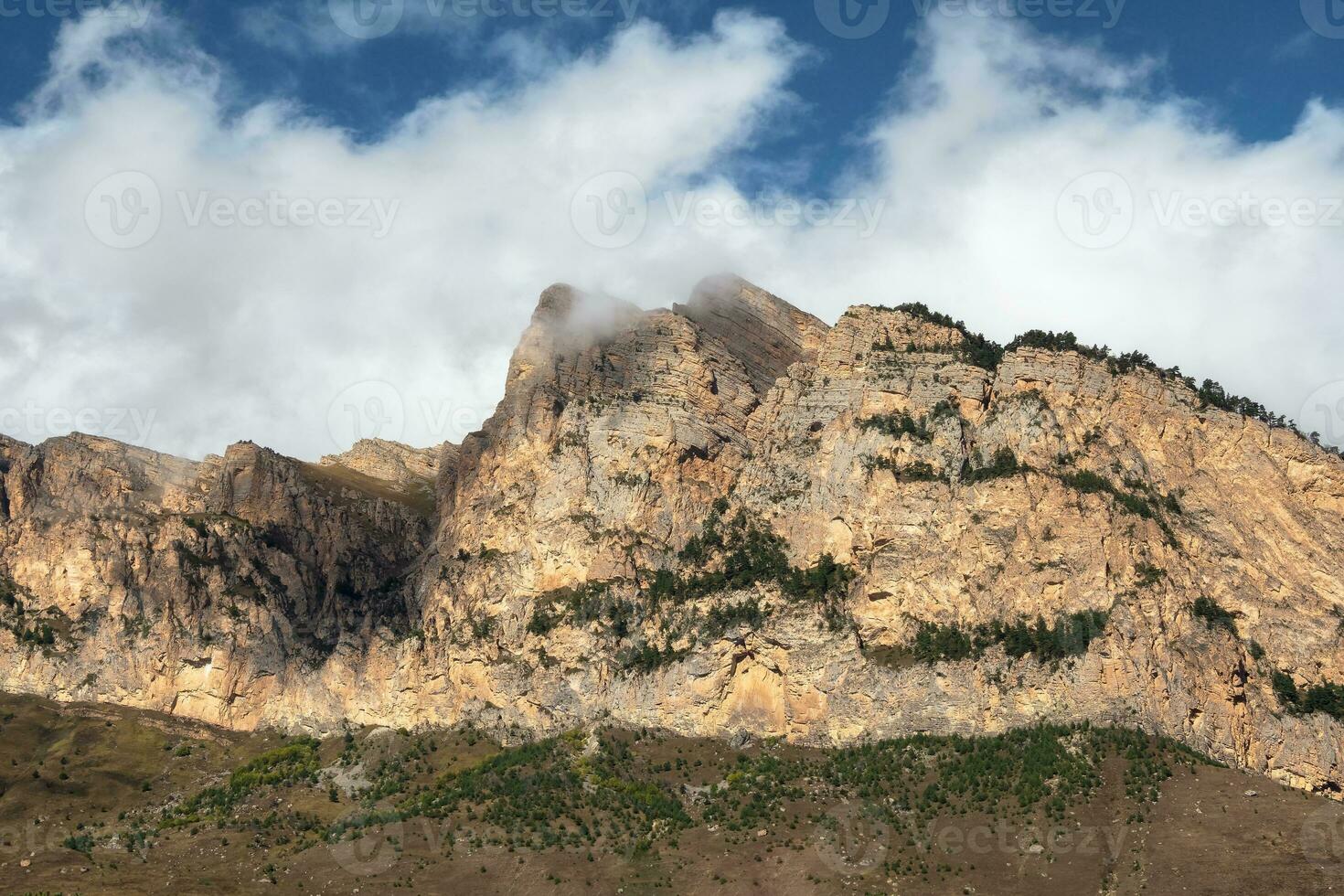 hermosa largo puntiagudo rock es iluminado por el Dom. soleado montaña paisaje con alto agudo Montañas Rocosas debajo blanco nubes panorámico ver de agudo rocas y paz parte superior en azul nublado cielo. foto