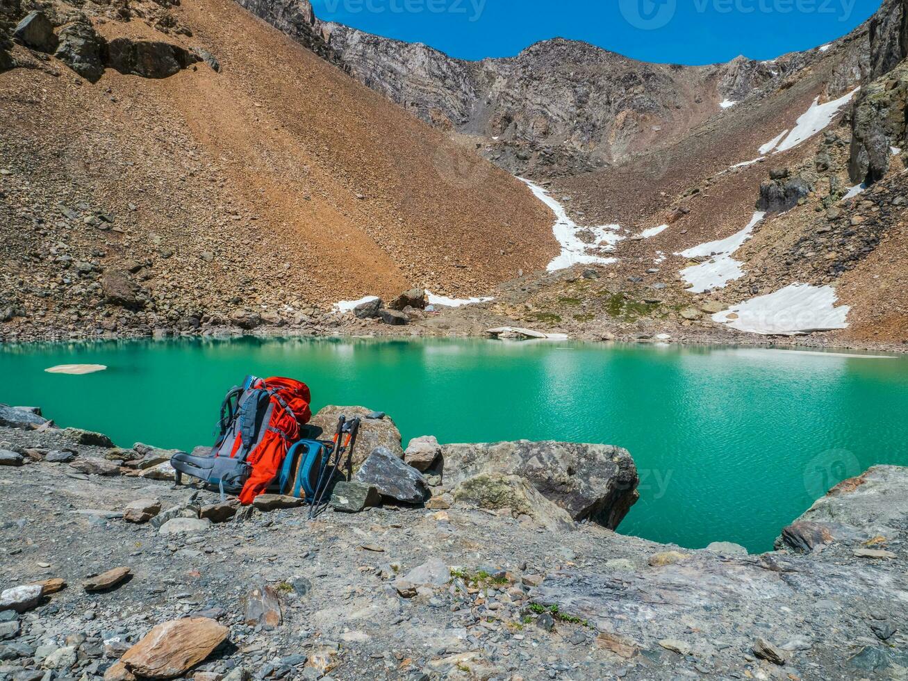 A large orange backpack and trekking poles on the shore of a mountain lake. Hiking in the highlands. The time of the stop on the hike. photo