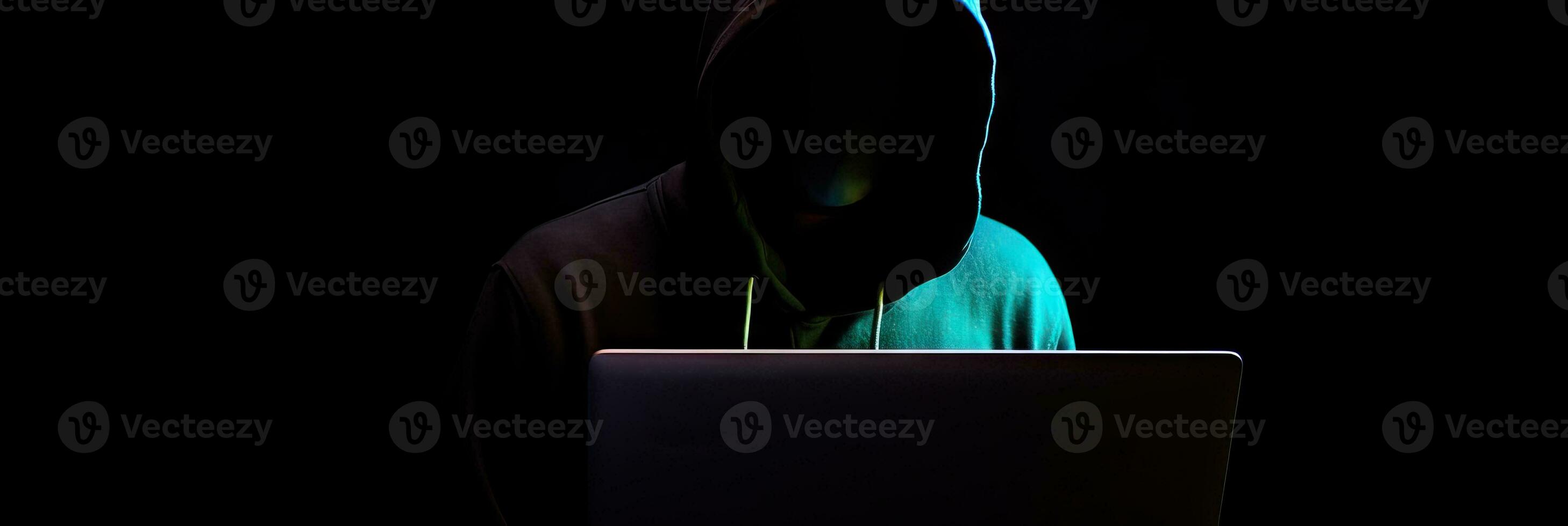 Hacker without face in a hood holds a laptop. on a dark background. Banner for cyber security.  Internet web hack technology. Digital laptop in hacker man hand isolated on black. photo