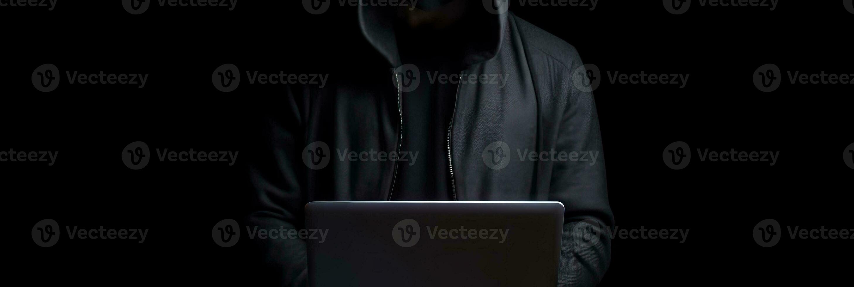 Hacker without face in a hood holds a laptop. on a dark background. Banner for cyber security.  Internet web hack technology. Digital laptop in hacker man hand isolated on black. photo