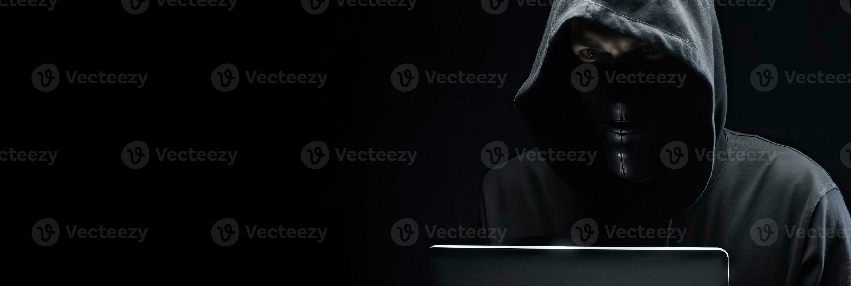 Hacker without face in a hood holds a laptop. on a dark background. Banner for cyber security.  Internet web hack technology. Digital laptop in hacker man hand isolated on black. photo