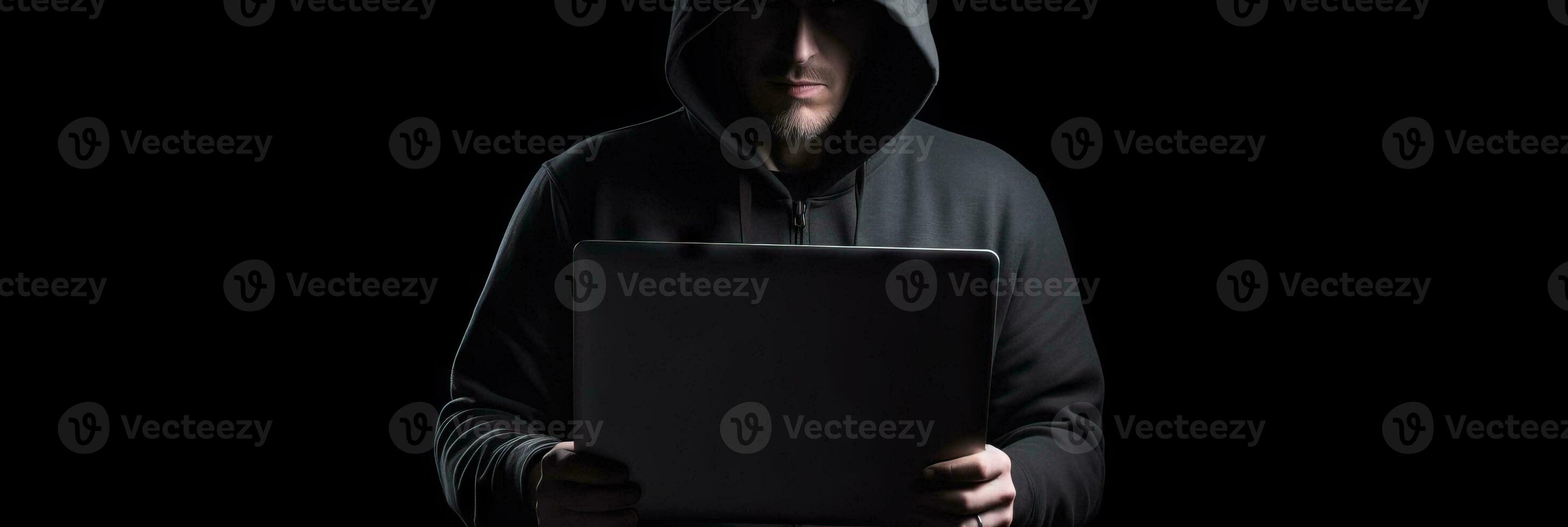 Hacker without face in a hood holds a laptop. on a dark background. Banner for cyber security.  Internet web hack technology. Digital laptop in hacker man hand isolated on black. photo