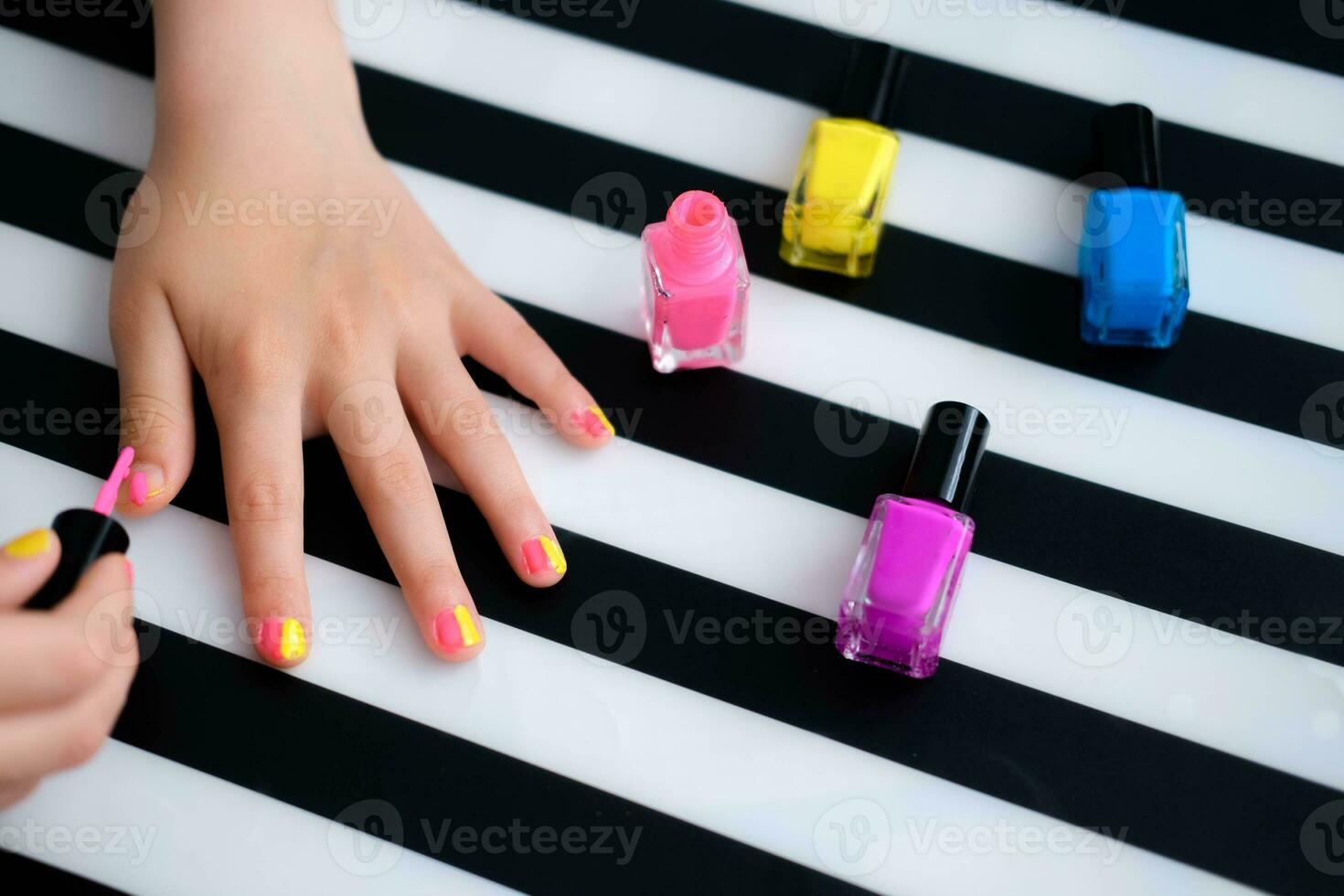 The child sits at the table and paints his nails with bright pink nail polish. A little girl does a manicure. Beauty salon game. Children is fun photo