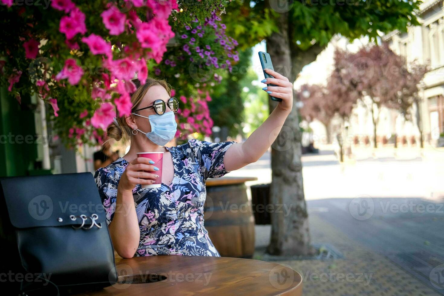 Beautiful young woman in a cafe in a sunglasses and medical mask and makes a selfie on the phone. A girl in a cafe on the street communicates online on a smartphone. Pandemic covid-19, quarantine. photo