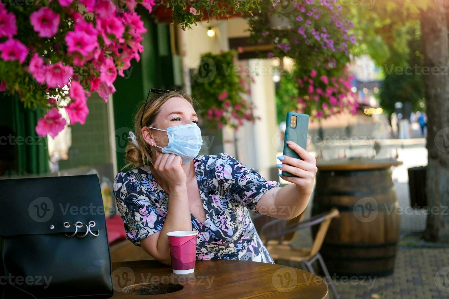 Beautiful young woman in a cafe in a medical mask and makes a selfie on the phone. A girl in a cafe on the street communicates online on a smartphone. Pandemic covid-19, quarantine. photo