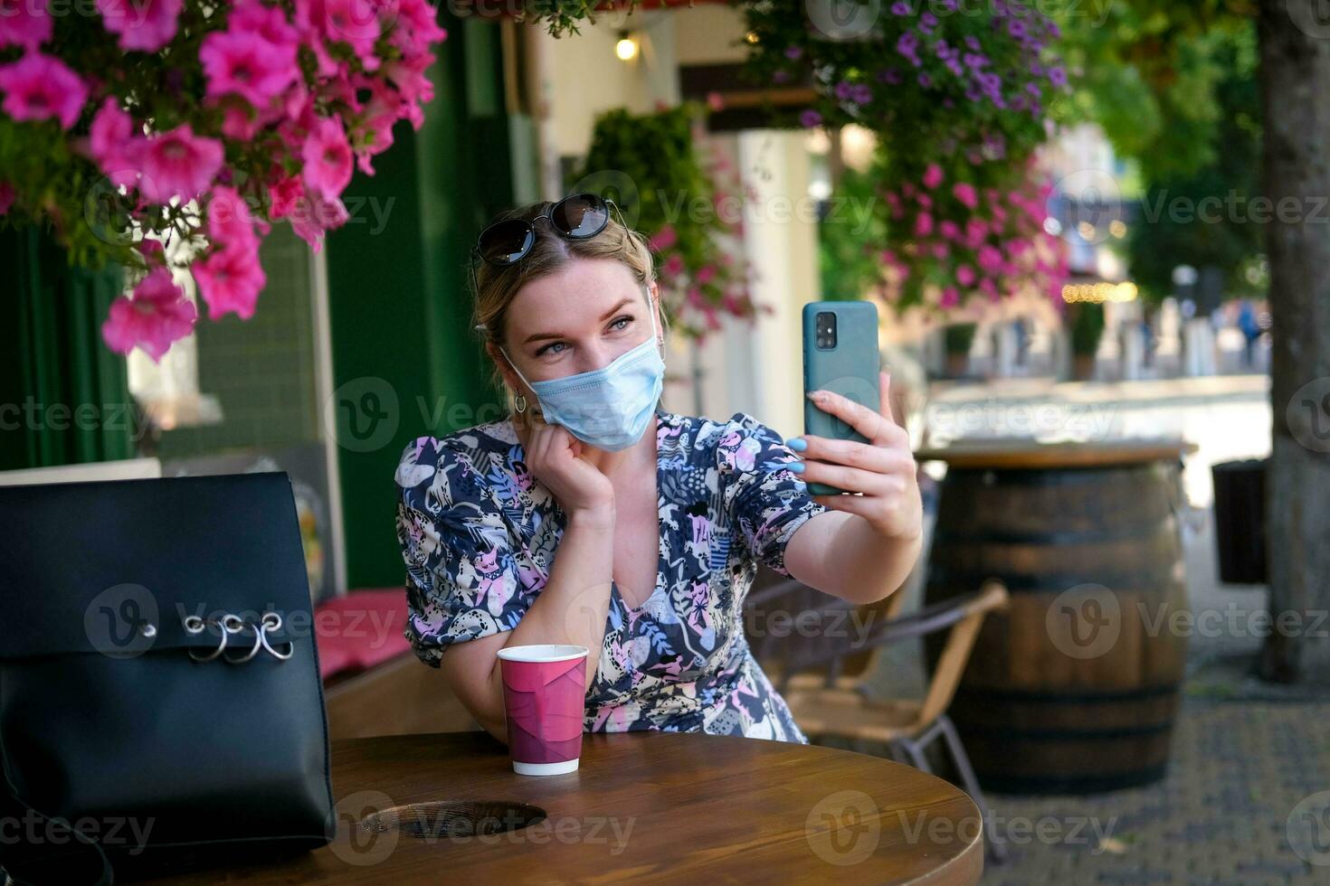 Beautiful young woman in a cafe in a medical mask and makes a selfie on the phone. A girl in a cafe on the street communicates online on a smartphone. Pandemic covid-19, quarantine. photo