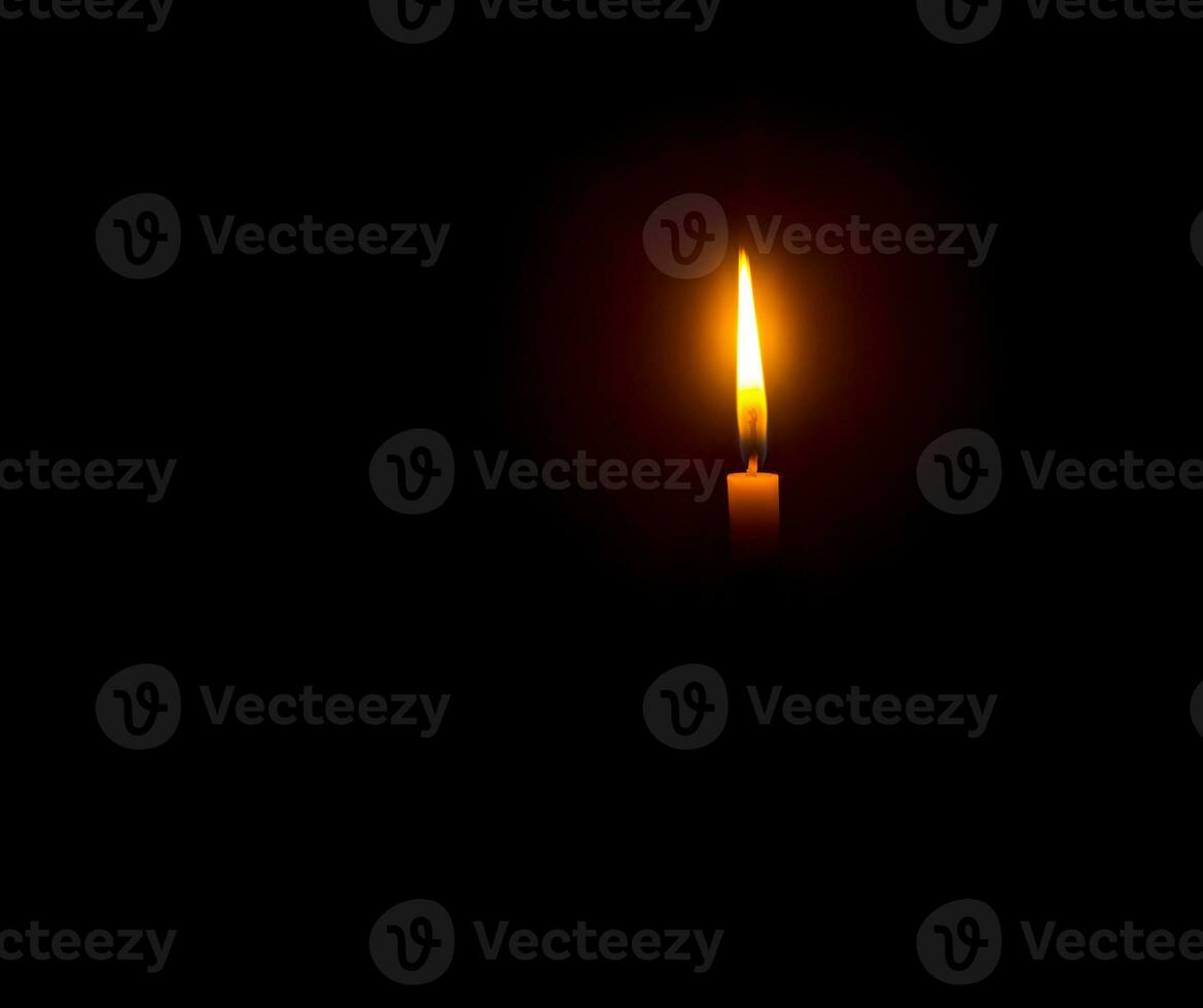 A single burning candle flame or light glowing on an orange candle on black or dark background on table in church for Christmas, funeral or memorial service with copy space photo