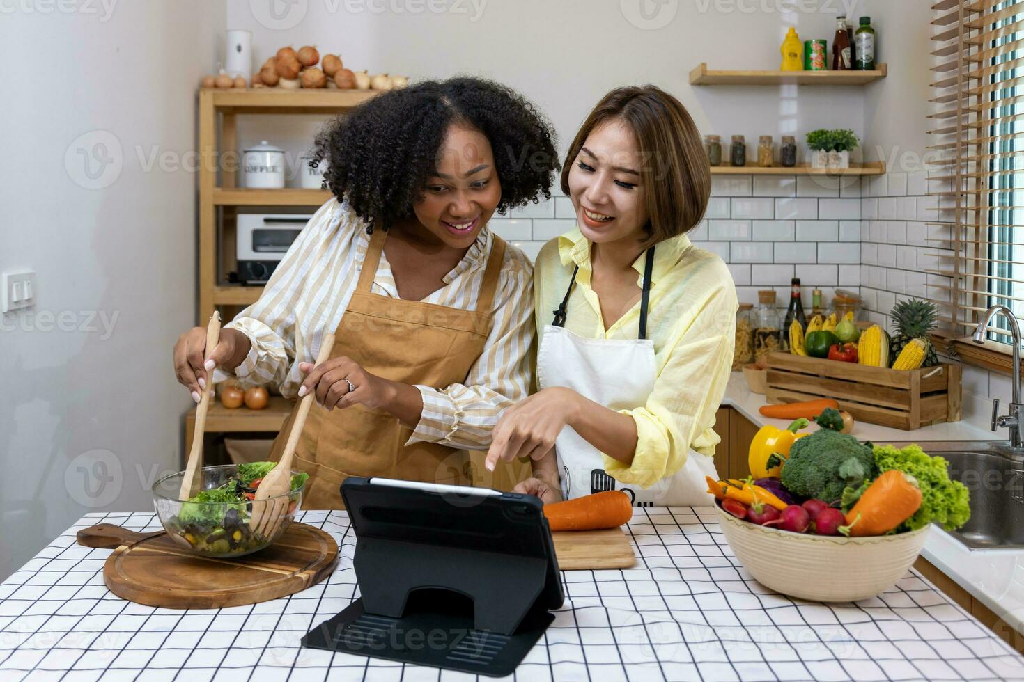 africano americano mujer y su asiático amigo es Ayudar cada otro preparando sano comida con variedad de vegetales mientras mirando a el Cocinando instrucción desde Internet utilizando digital tableta foto