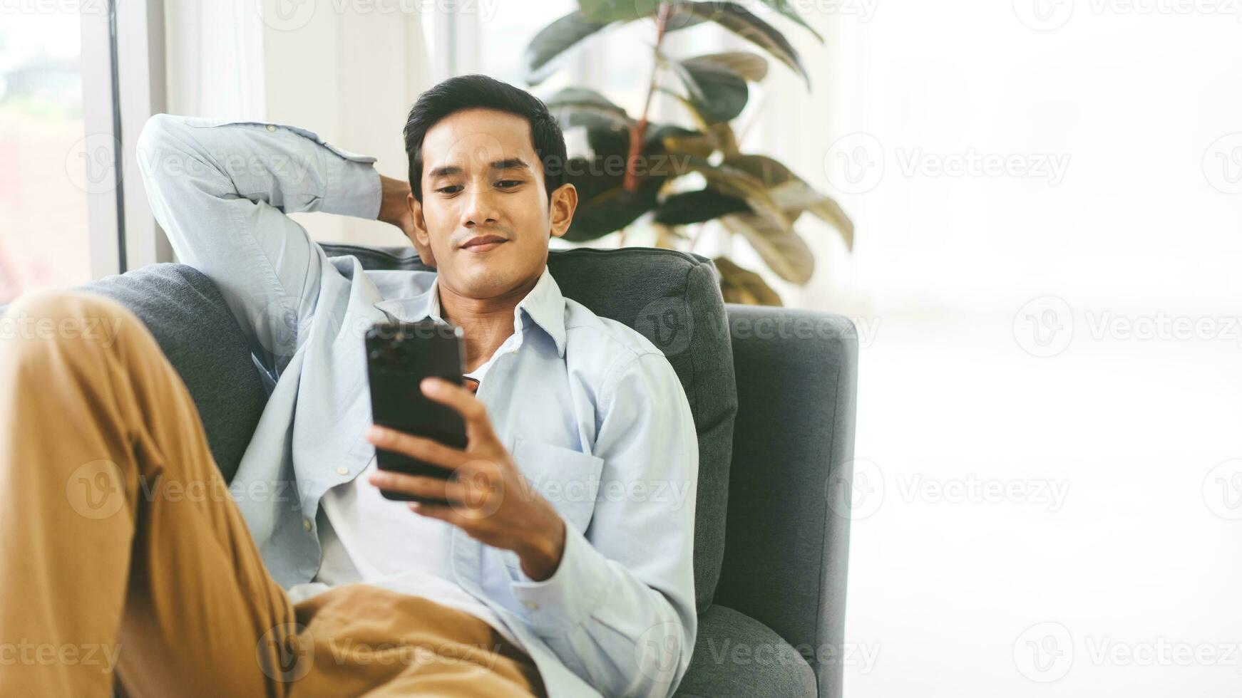 Young adult southeast asian man relax on sofa using smartphone for online leisure at home background with copy space photo