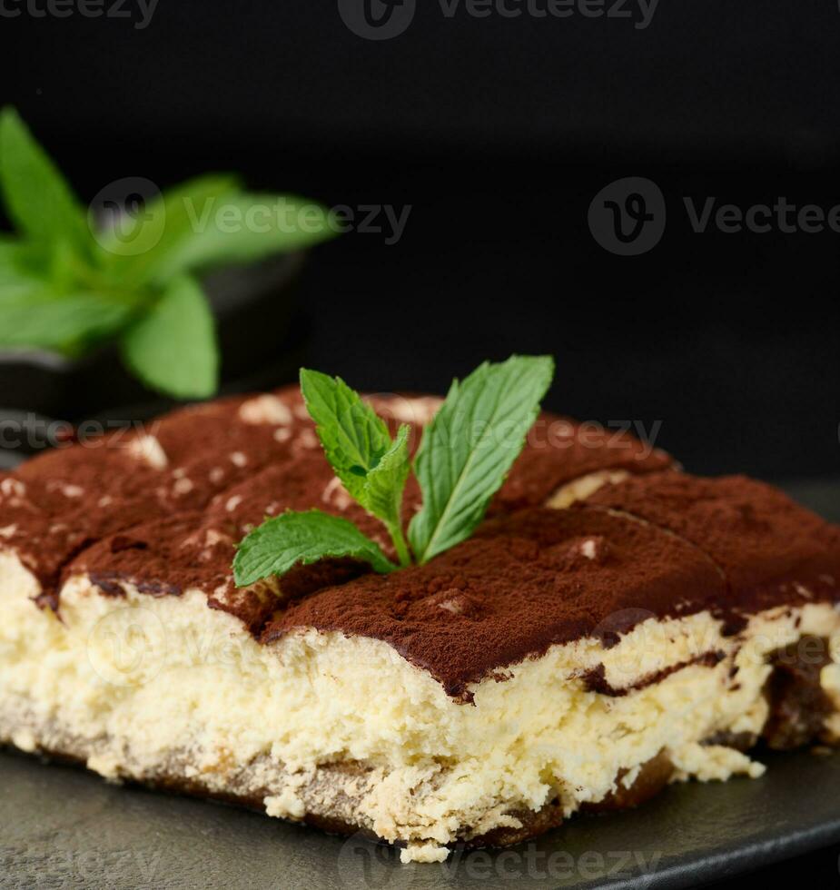 A piece of tiramisu sprinkled with cocoa, on top of a sprig of fresh mint on a black background photo