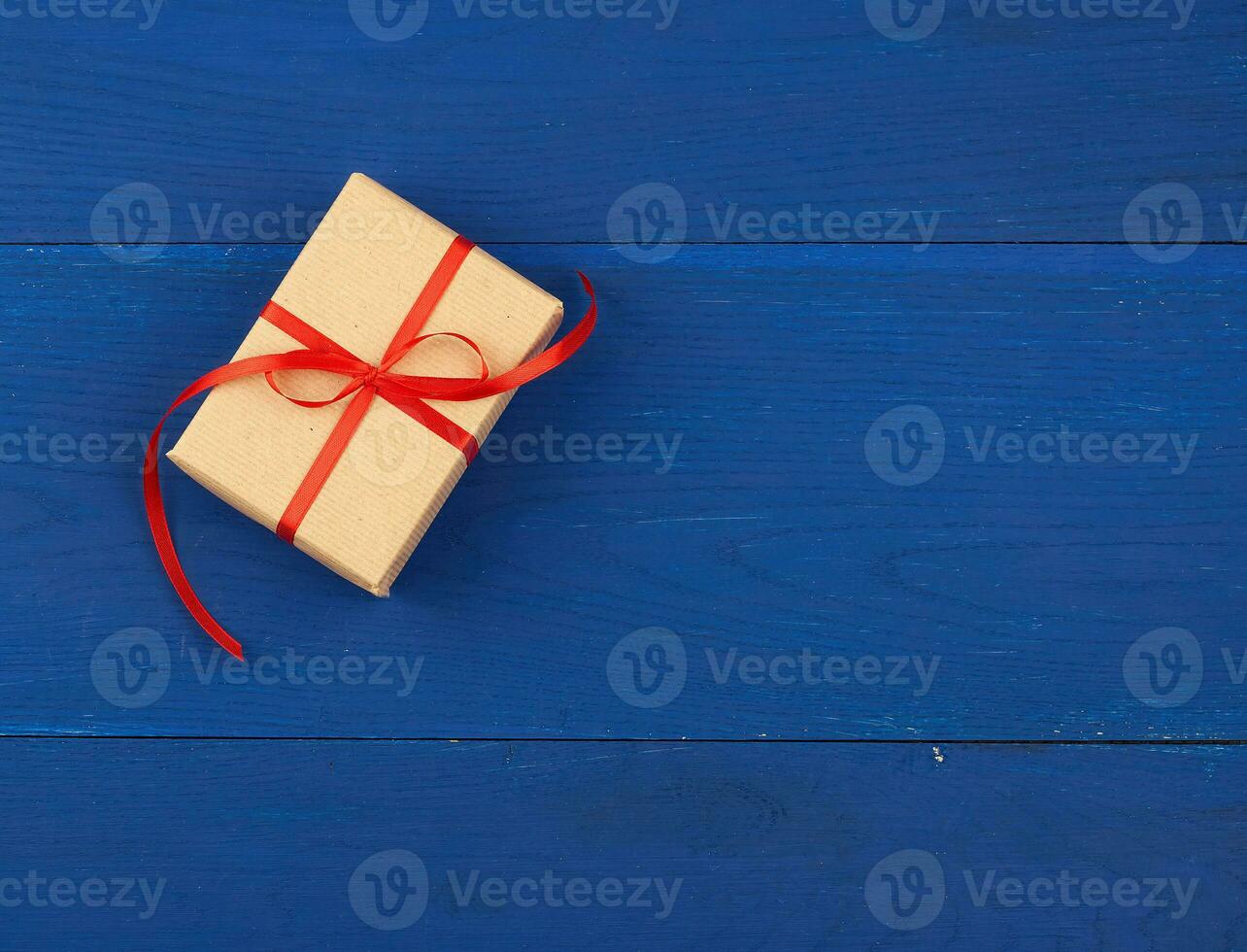 gift wrapped in brown kraft paper and tied with a thin red ribbon on a blue wooden background, top view, flat lay. Eco package design photo