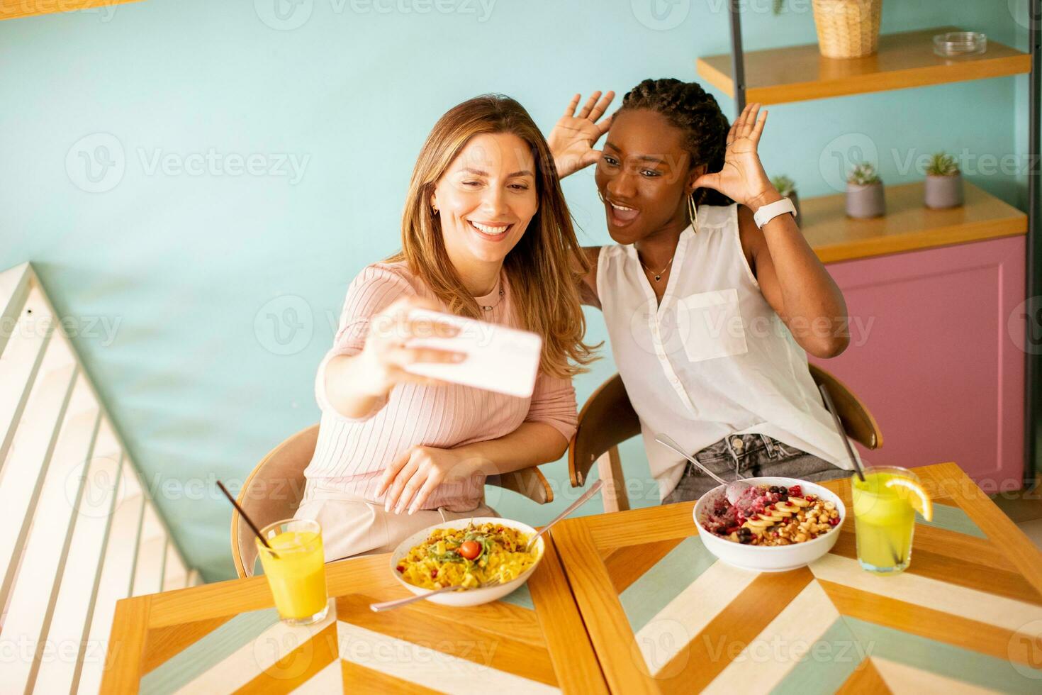 Two young women, caucasian and black one, taking selfie with mobile phone in the cafe photo