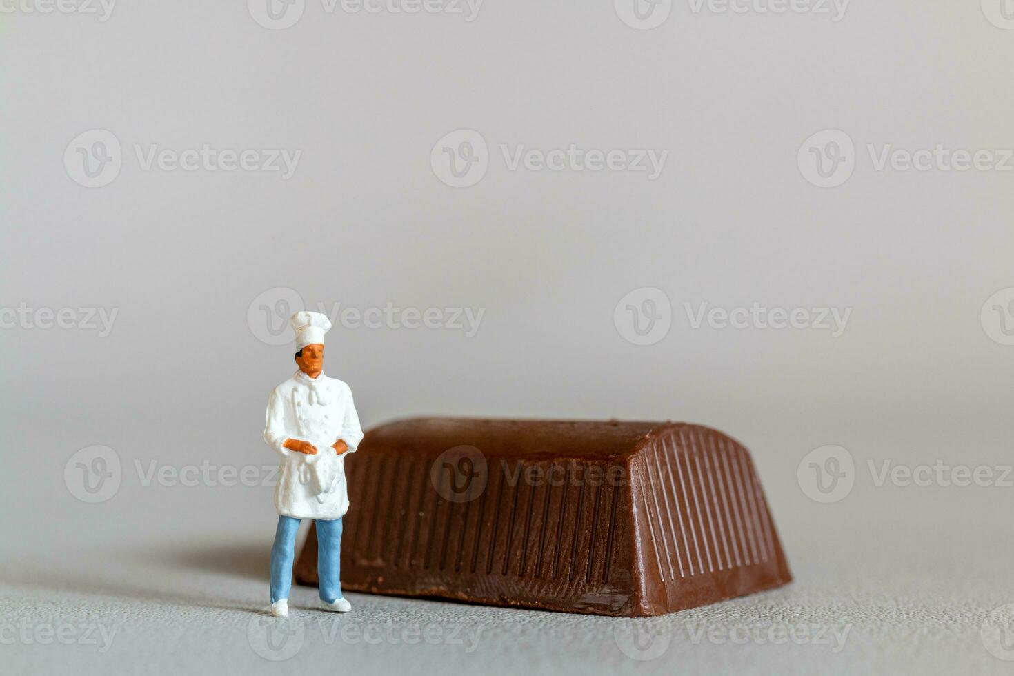 miniatura personas cocinero con chocolate en pie mientras en pie en contra un gris antecedentes foto