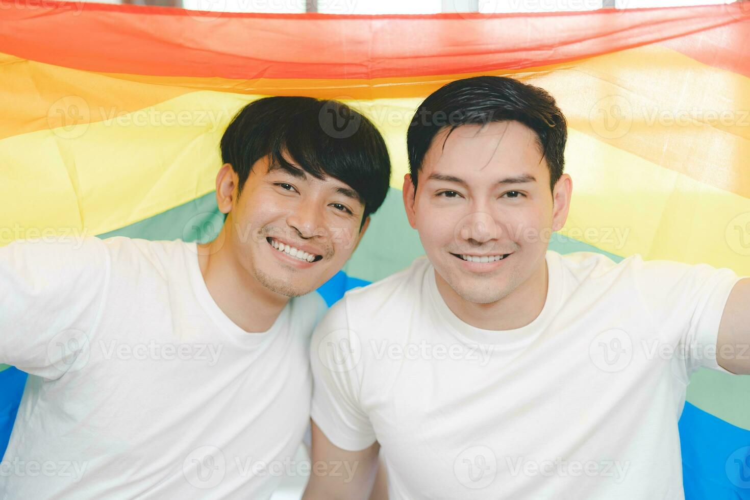 Happy Asian LGBT couple, sitting on the sofa holding and waving rainbow LGBT Pride flag together in the bedroom at home. Diversity of LGBT relationships. A gay couple concept. Loving gay couple. photo