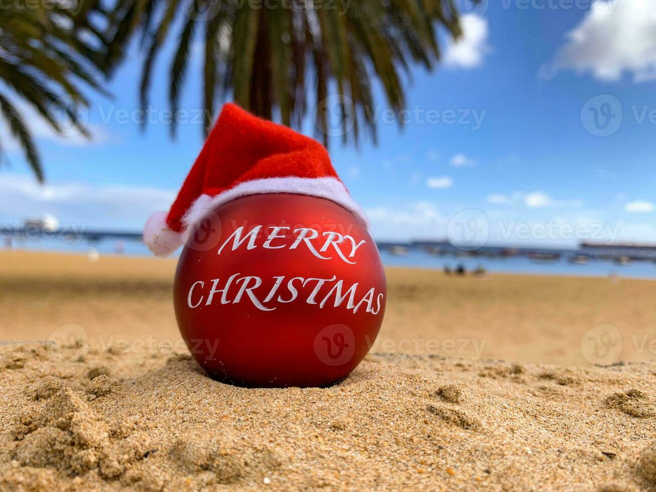 Christmas bomb in Santa's hat on the beach lying on the sand with palm trees and blue sky on the background. Merry christmas from paradise, exotic island. Hawaii, Canary islands, Bali. photo