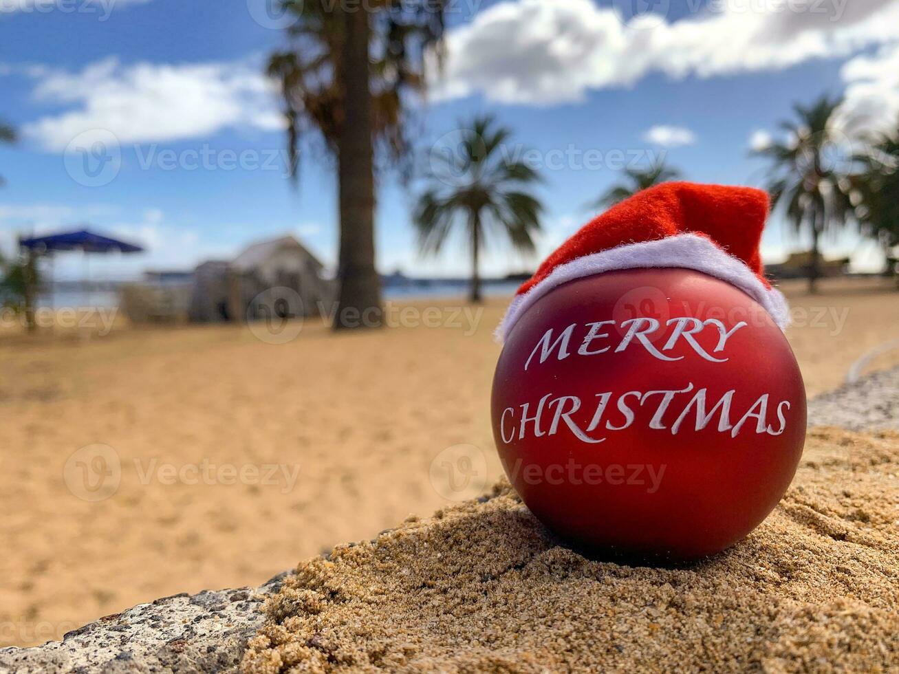 Christmas bomb in Santa's hat on the beach lying on the sand with palm trees and blue sky on the background. Merry christmas from paradise, exotic island. Hawaii, Canary islands, Bali. photo