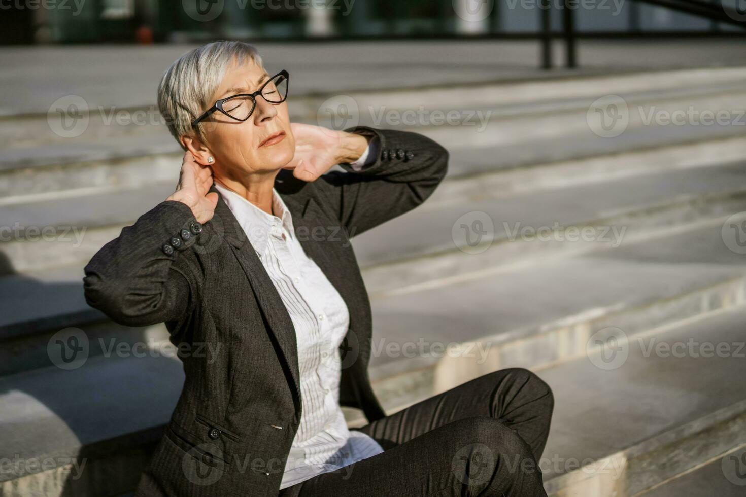 Mature businesswoman sitting in front of company building. She is tired and having pain in neck. photo