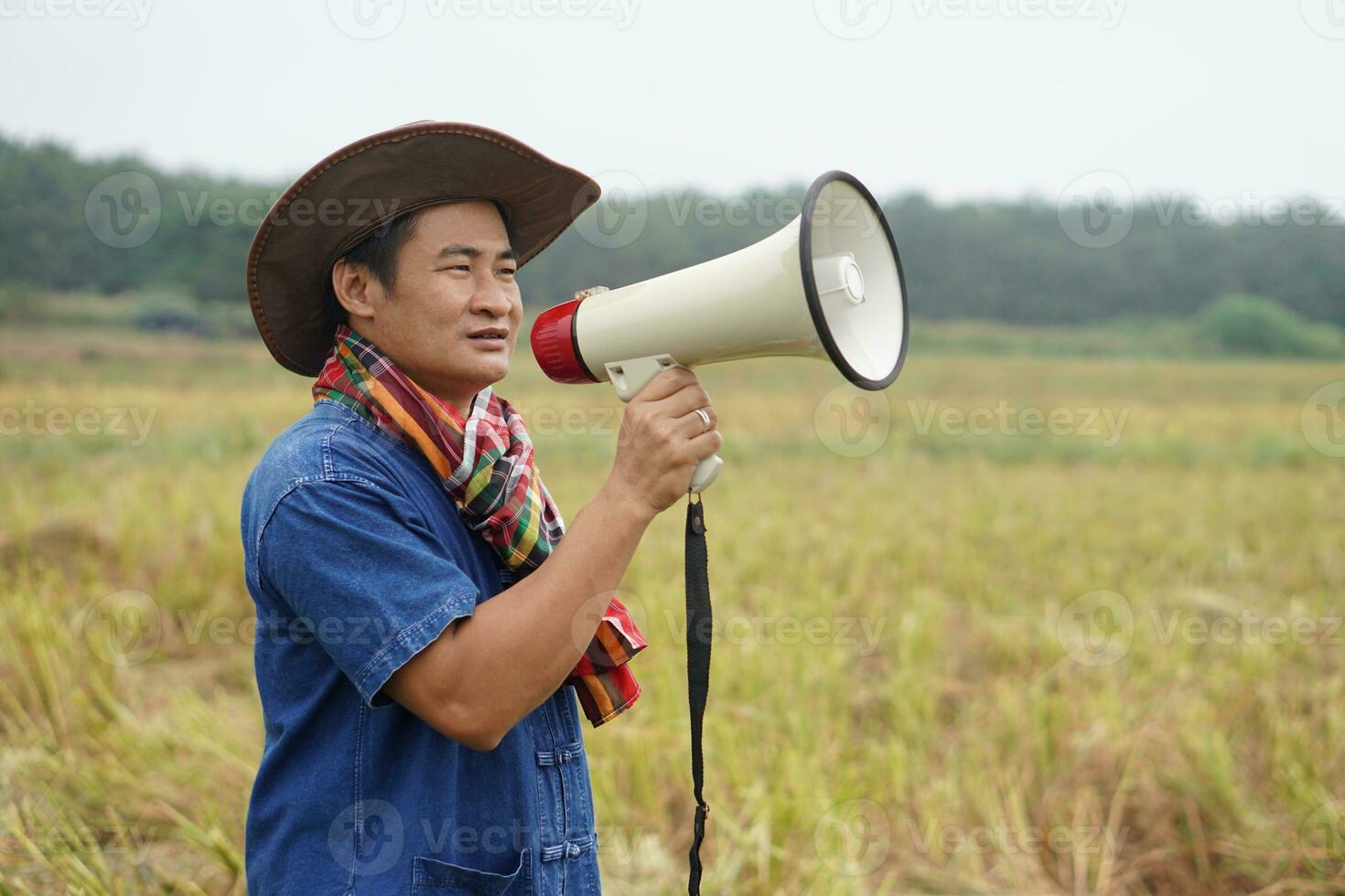 Asian man farmer wears hat, holds megaphone at agriculture field. Concept, communication , announce. Protest. Protester. Bullhorn public address. Loudspeaker technology for outdoor announcement. photo
