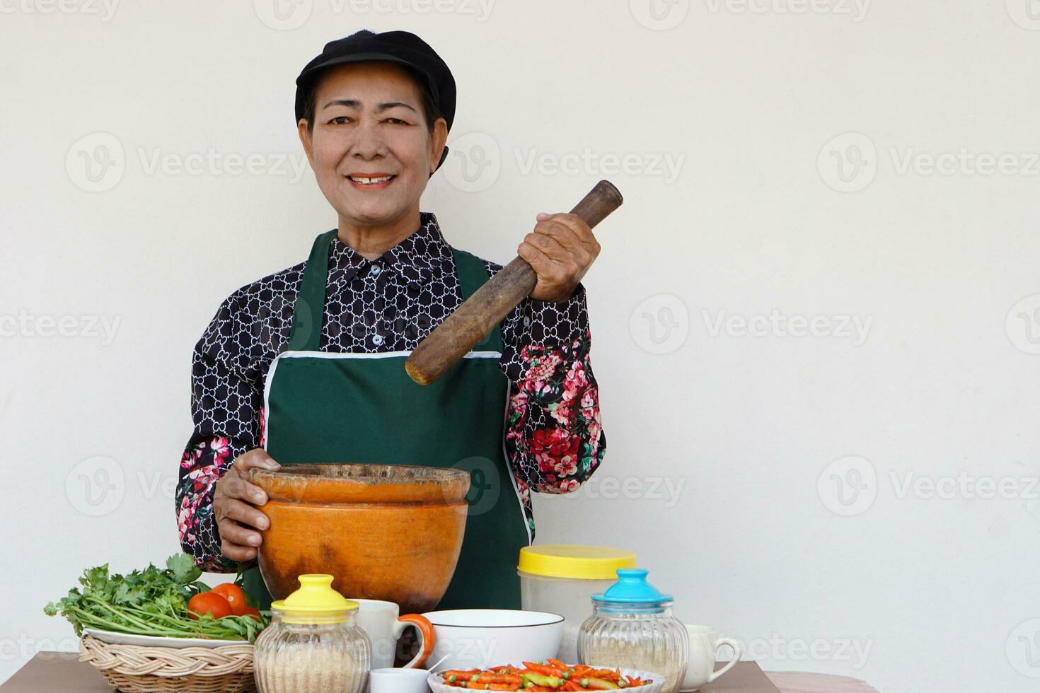 Happy Asian senior woman is cooking, wear chef cap and apron, holds pestle, mortar and plate of chillies. Concept, Cooking for family. Thai kitchen lifestyle. Elderly activity. photo