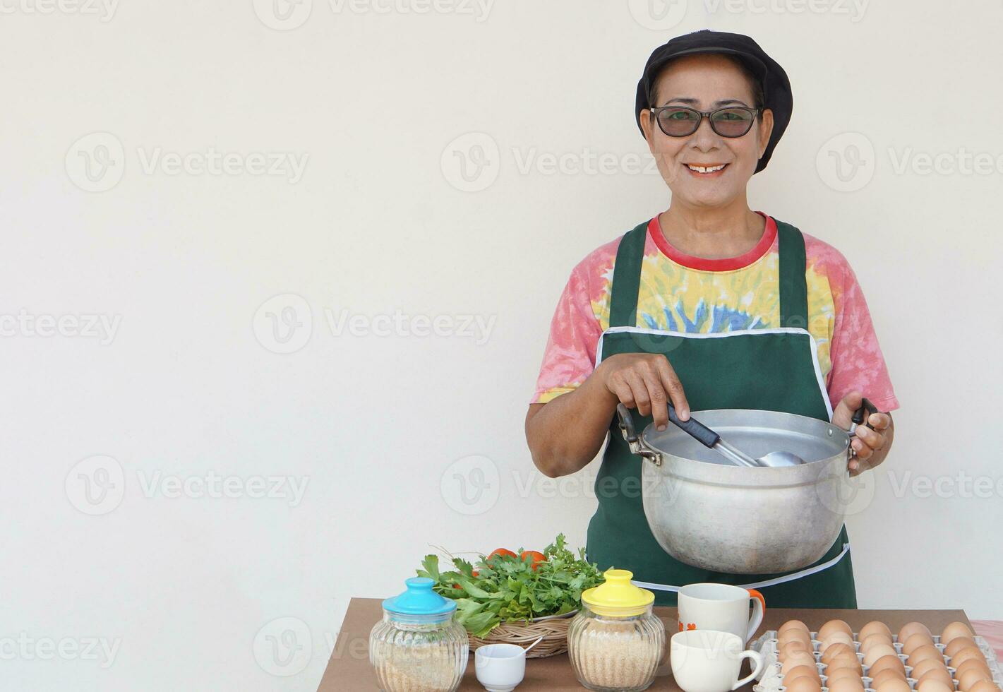 contento asiático mayor mujer es cocinando, vestir cocinero gorra y delantal, sostiene maceta y cucharón. concepto, Cocinando para familia. Cocinando para vender. tailandés cocina estilo de vida. mayor actividad. foto