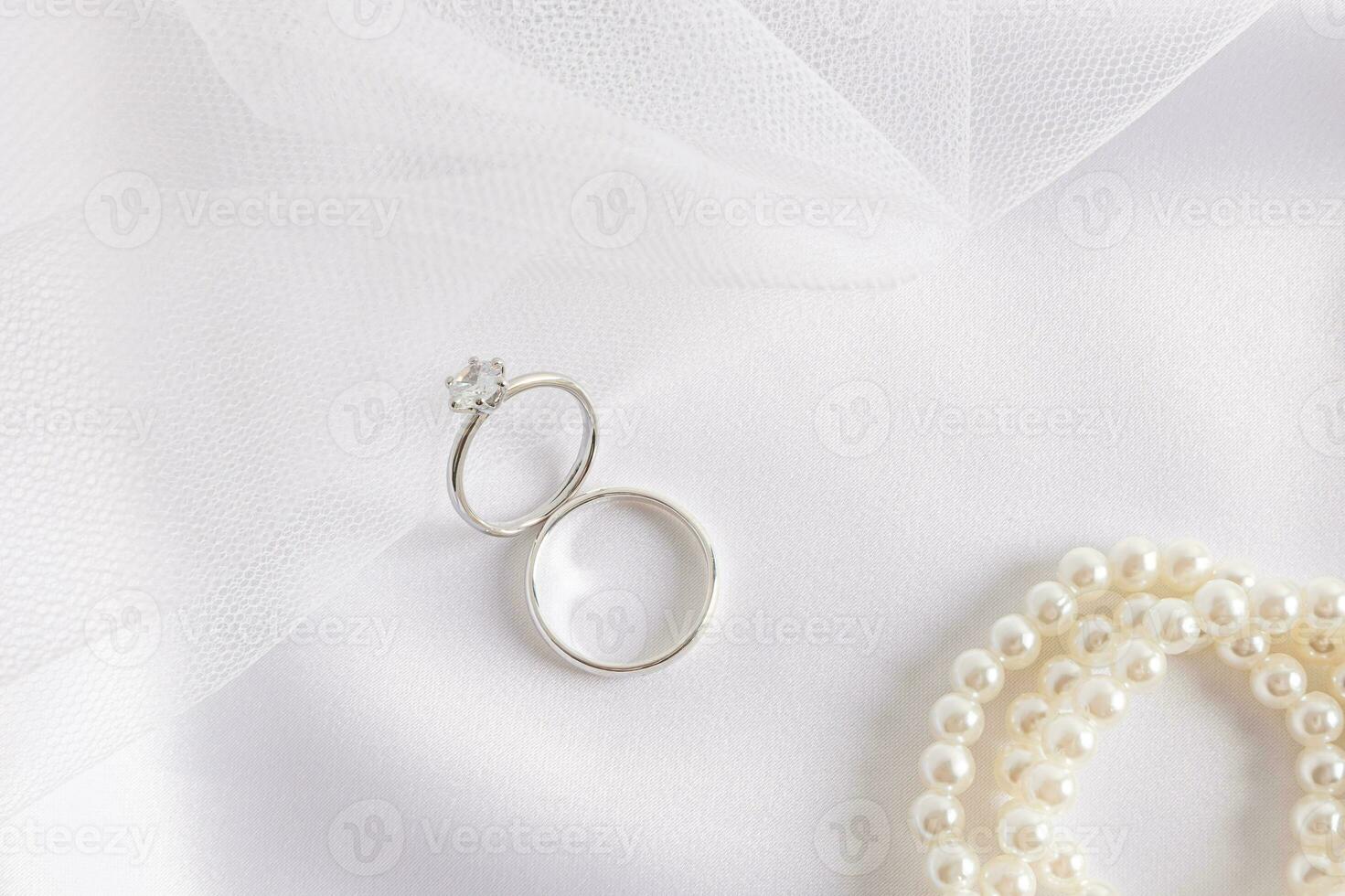 Two white gold rings with a diamond on the part of the bride's wedding veil and satin white textiles with pearl beads. Wedding composition. photo